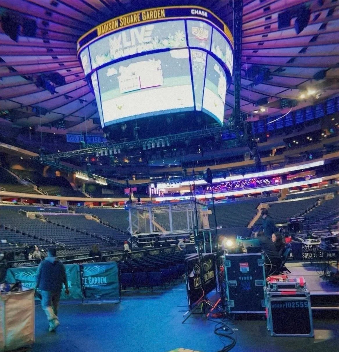 Empty indoor arena with a large circular LED screen displaying live event graphics, set up for a concert or event at Madison Square Garden, with some crew members managing equipment on the stage for WWE Holiday Tour.
