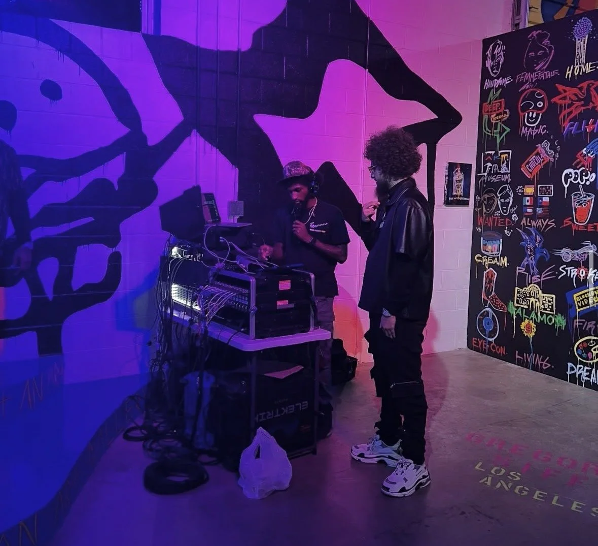 Two men in a colorful, neon-lit art space with collage and graffiti on the walls. One man is DJing with equipment, and the other is wearing a leather jacket and sneakers, engaging in conversation.