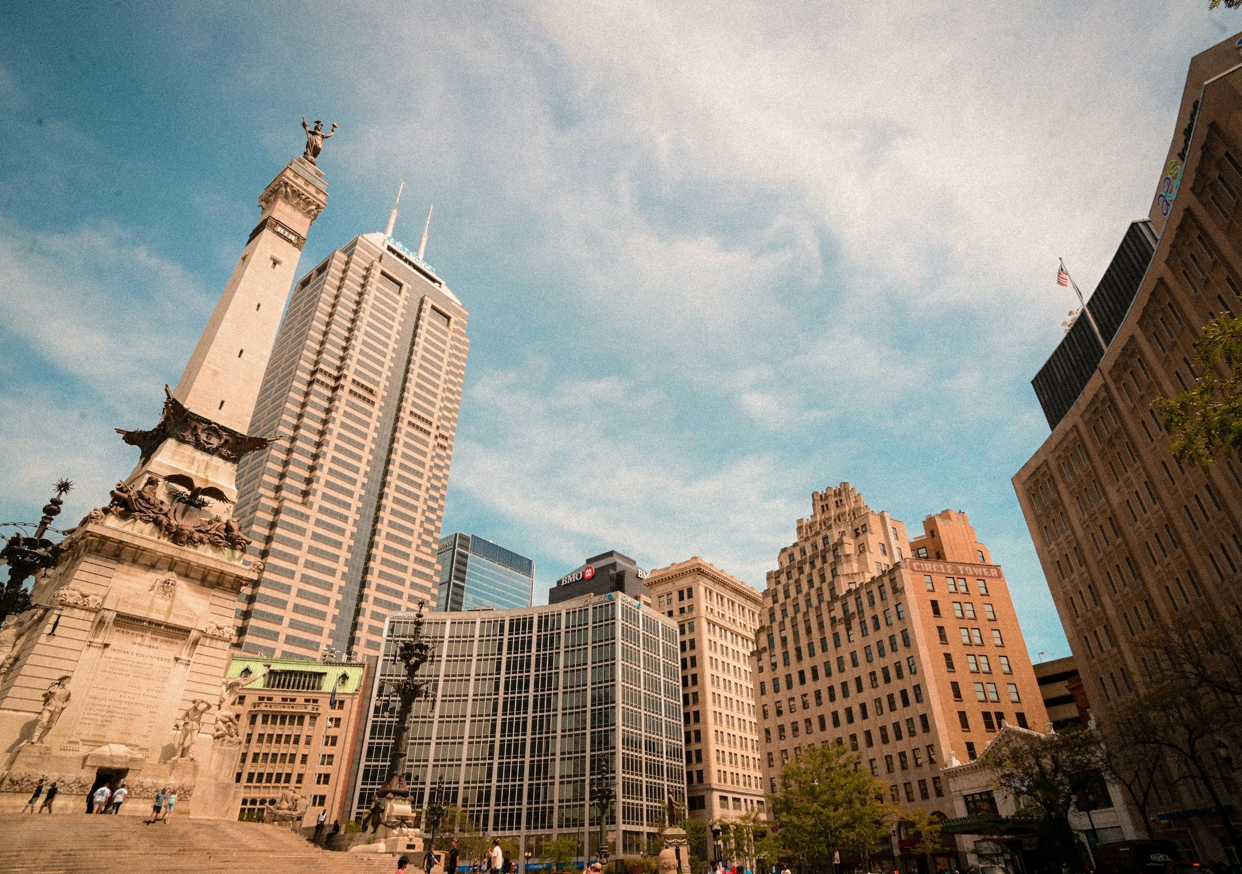 View of a city square with tall skyscrapers and a monument featuring a statue on top. There are people walking around and the sky is partly cloudy.