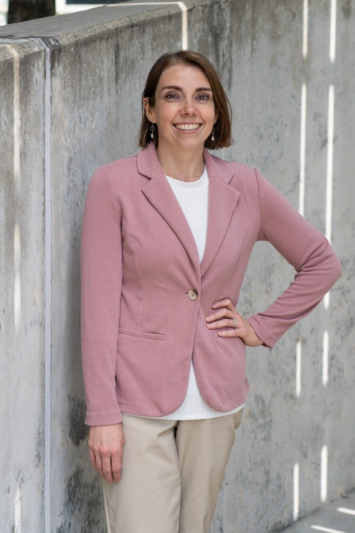 A woman with short brown hair, wearing earrings, a pink blazer, a white top, and beige pants, standing outdoors against a concrete wall, smiling and with one hand on her hip.