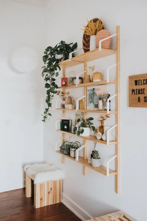 Wooden wall-mounted shelf with plants, books, and decorative items, next to a small bench with a fur cushion, in a minimalist room with white walls and wooden flooring.