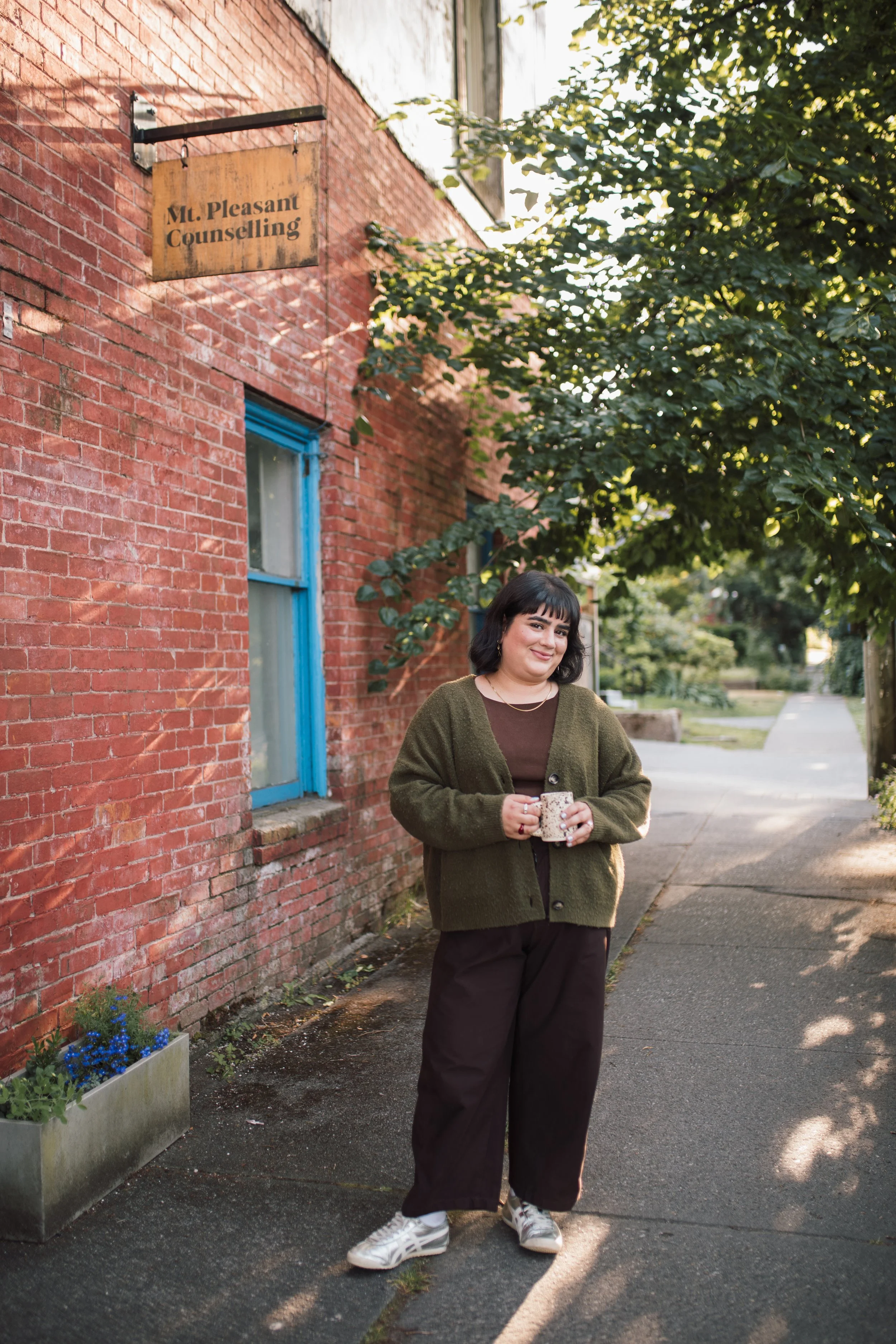 A woman standing outdoors on a sidewalk with a brick building and green trees behind her, holding a mug and smiling.