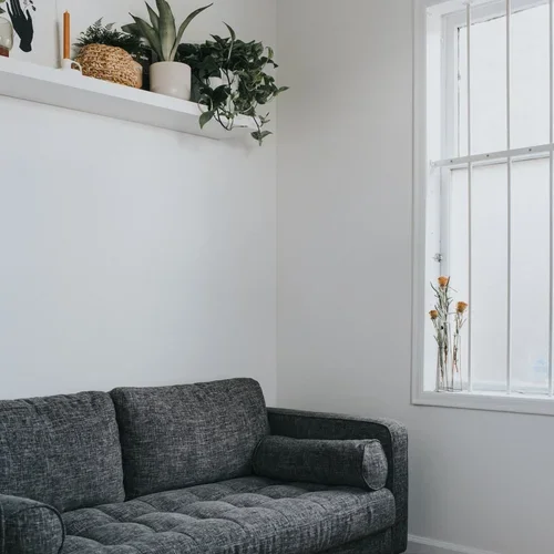 A gray couch in a bright, minimalistic living room with a window and a white shelf with plants and decor.