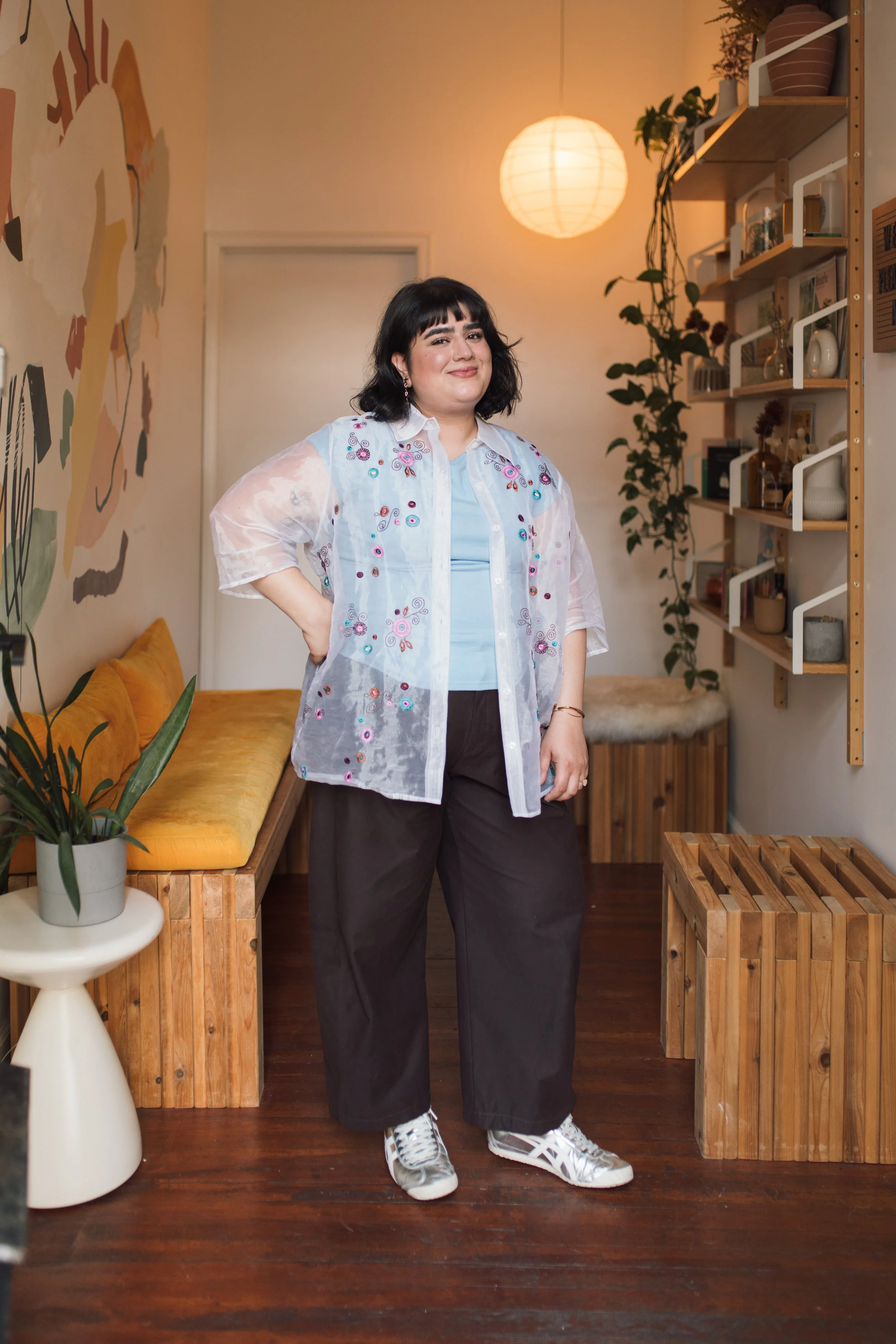 A woman with shoulder-length black hair and bangs stands in a warmly lit room with wooden furniture and plants. She is wearing a light blue shirt under a sheer, embroidered jacket and dark pants, with her right hand on her hip and smiling slightly. The room has a yellow couch, a floor lamp, shelves with decorative items, and a wall art.
