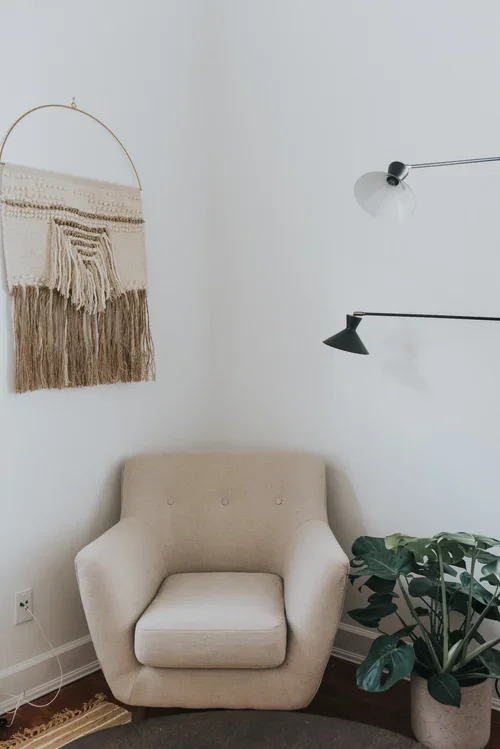 A cozy corner with a beige armchair, green houseplant, wall-mounted black lamp, and woven wall hanging with fringes on a white wall.