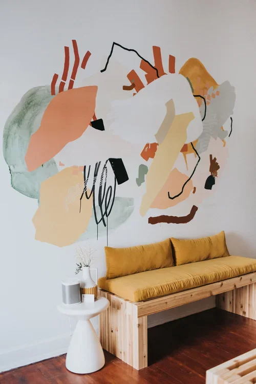 Abstract wall art with various shapes and colors above a mustard-yellow cushioned wooden bench in a room.