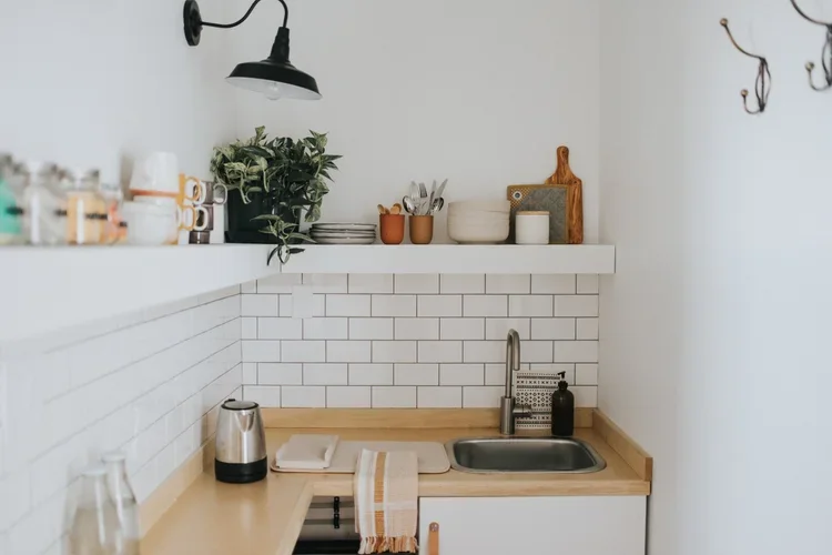 Small kitchen corner with a black wall-mounted lamp, a potted plant, stacked plates, cups, and cutting boards on a white shelf, white tiled backsplash, wooden countertop with a stainless steel sink, soap dispenser, and a yellow striped towel.