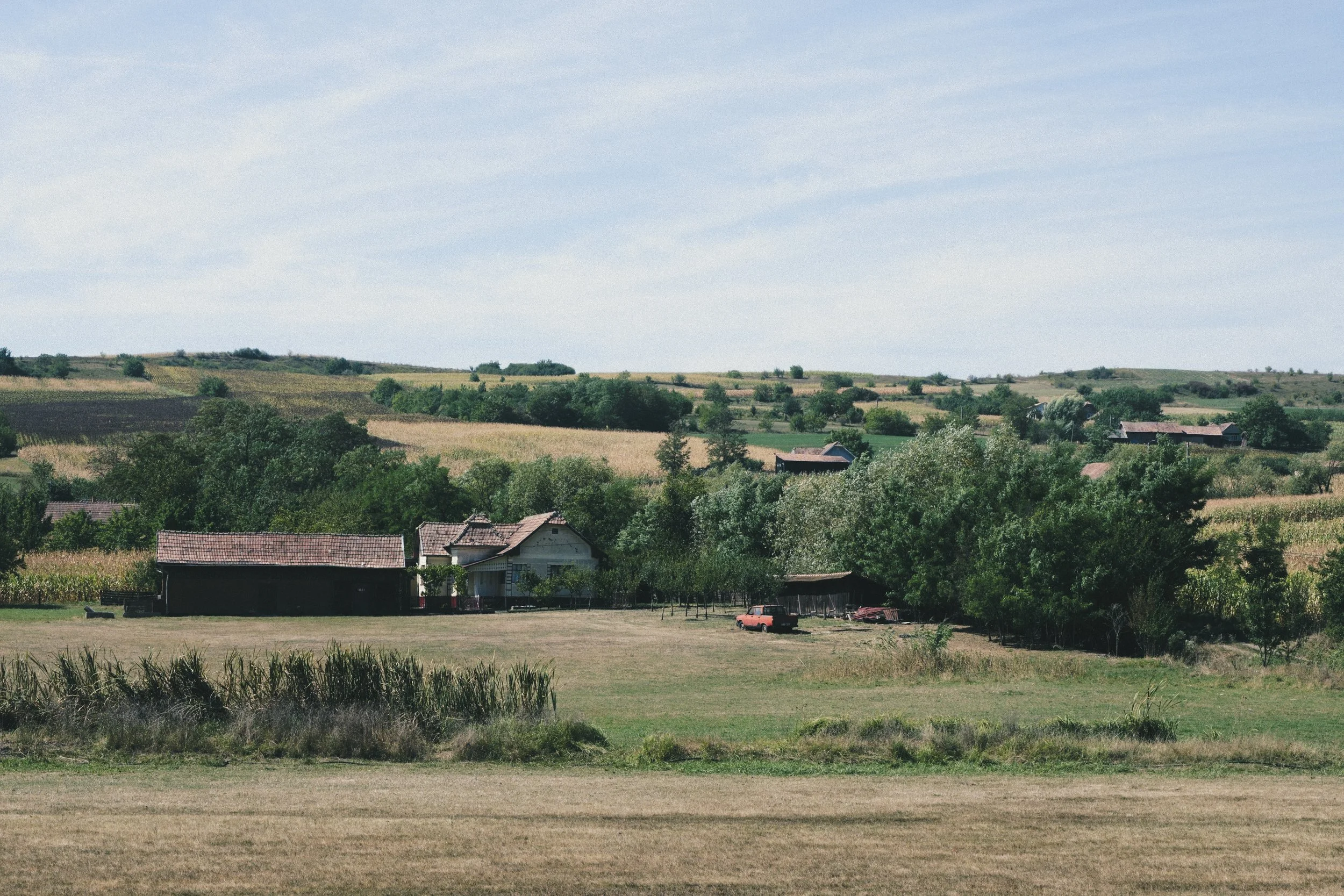 A rural landscape with rolling hills, scattered trees, farmhouses, and a small red vehicle near a fenced area under a cloudy sky.