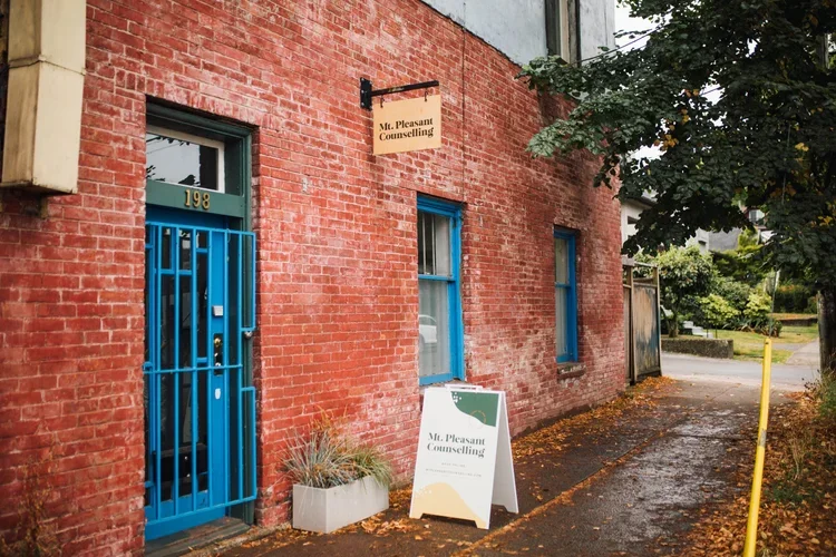 Red brick building with blue door and windows, with a sign for 'Mt. Pleasant Counselling' hanging above. A sidewalk with fallen leaves and a tree on the right side. An A-frame sidewalk sign for 'Mt. Pleasant Counselling' in front of the building.
