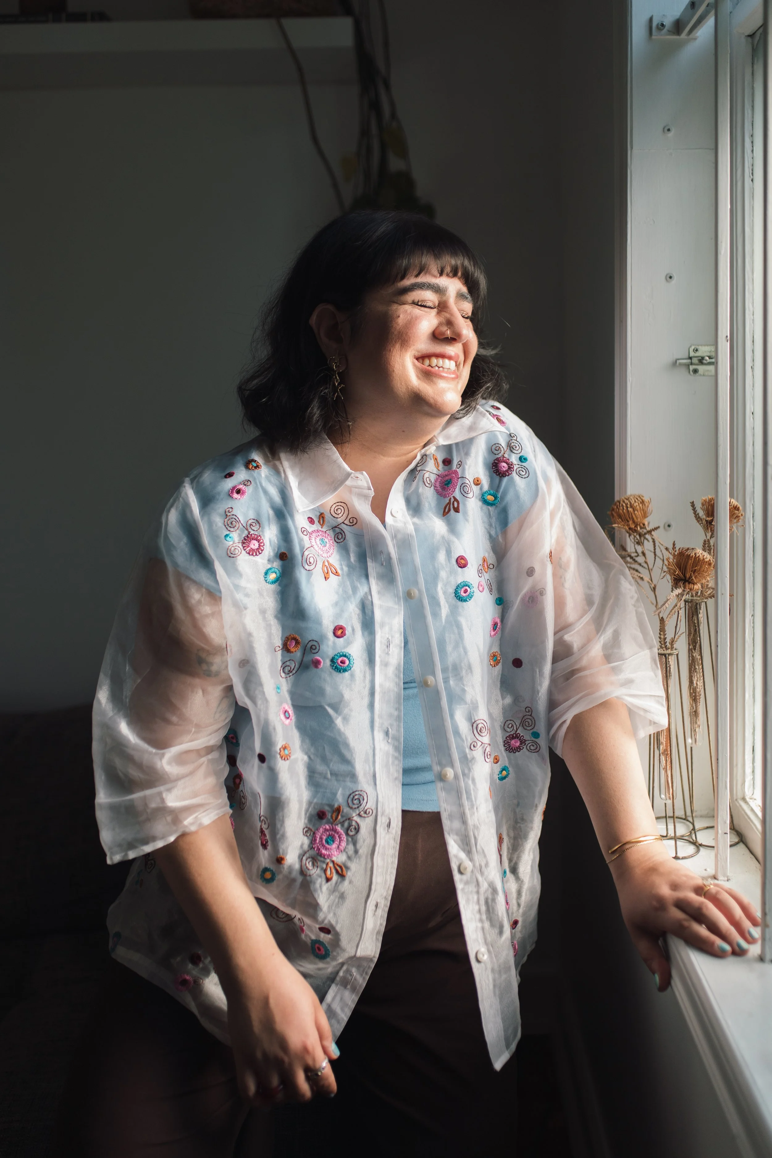 A woman with dark hair, smiling and closing her eyes, sitting by a window with natural light coming in. She wears a white embroidered shirt with colorful floral patterns.