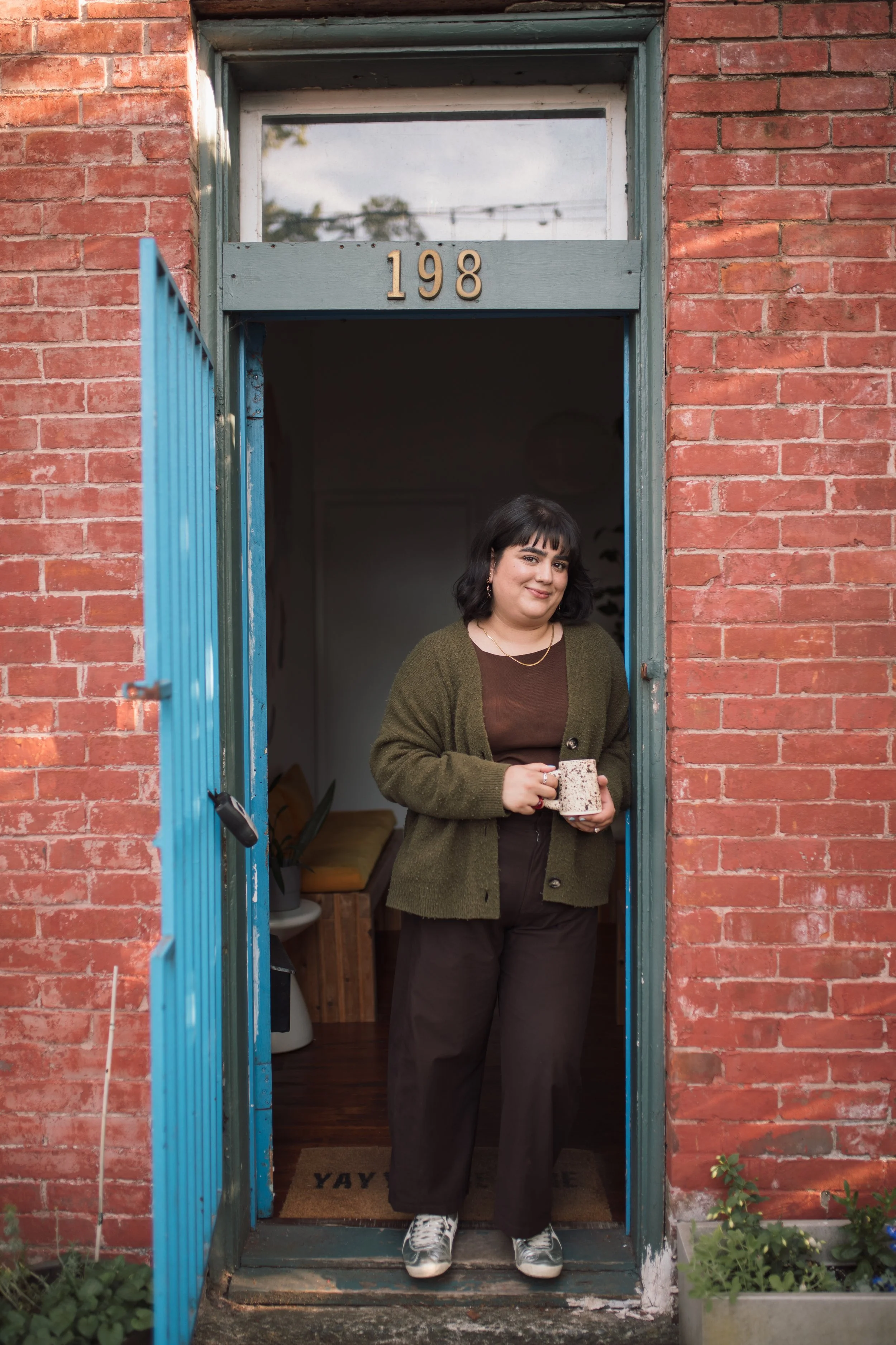 A woman with short black hair and bangs stands in an open doorway of a brick building, holding a mug in her hands and smiling at the camera. She is wearing a green cardigan over a brown top and black pants, with sneakers. The door has the number 198 above it, and there is a welcome doormat inside that says 'YAY!'. Plants are visible outside on either side of the doorway.