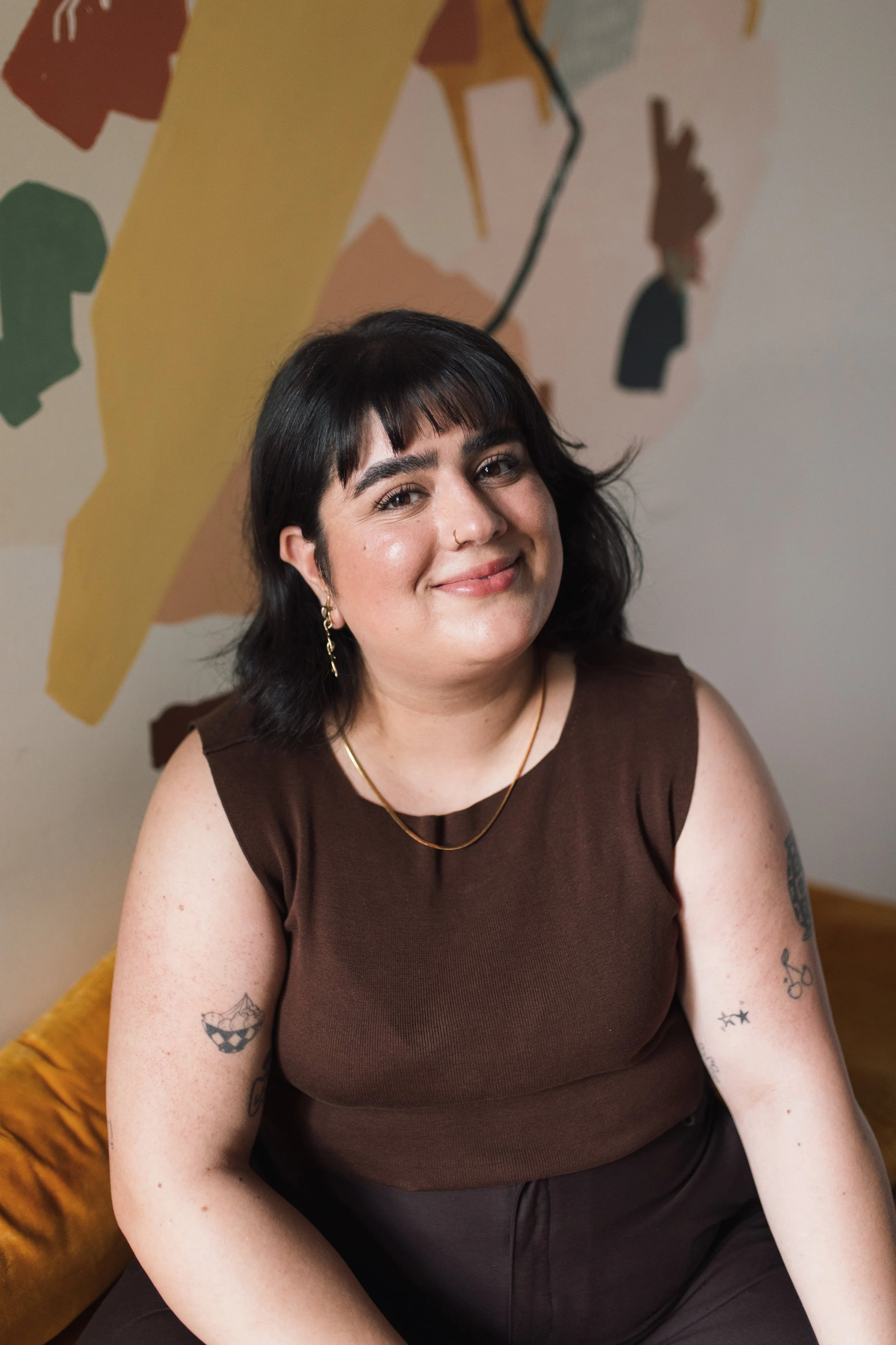 A woman with black hair, wearing a brown sleeveless top, sitting indoors in front of an abstract colorful mural. She is smiling and has tattoos on her left arm.