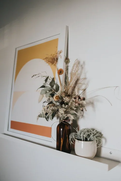 Decorative dried flower arrangement in a dark amber vase and a potted air plant on a white surface, with abstract artwork featuring warm colors hanging on the white wall.