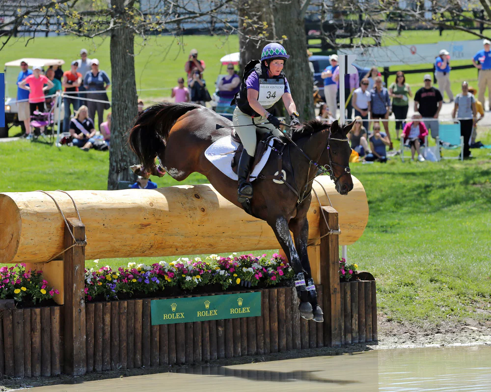 A female equestrian and her horse jumping over a water obstacle during an eventing competition, with spectators watching in the background.