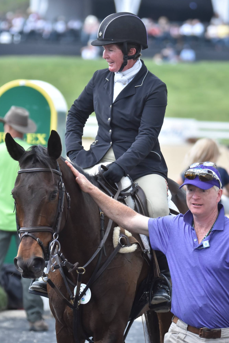 A woman riding a horse at a horse racing event, wearing a black riding helmet and dark jacket, smiling while another man in a purple shirt and sunglasses holds the horse gently.
