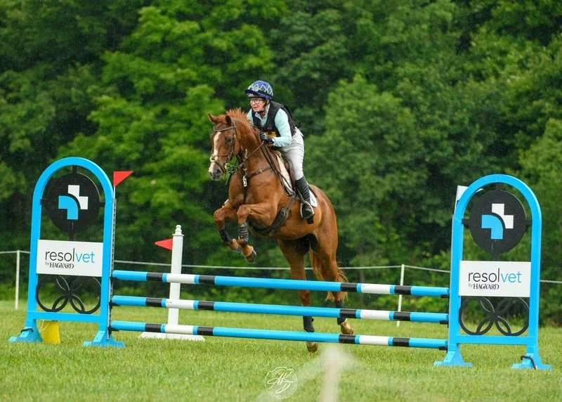 Sally Cousins and her horse jump over a blue and white obstacle during an equestrian event on a grassy field, with green trees in the background.