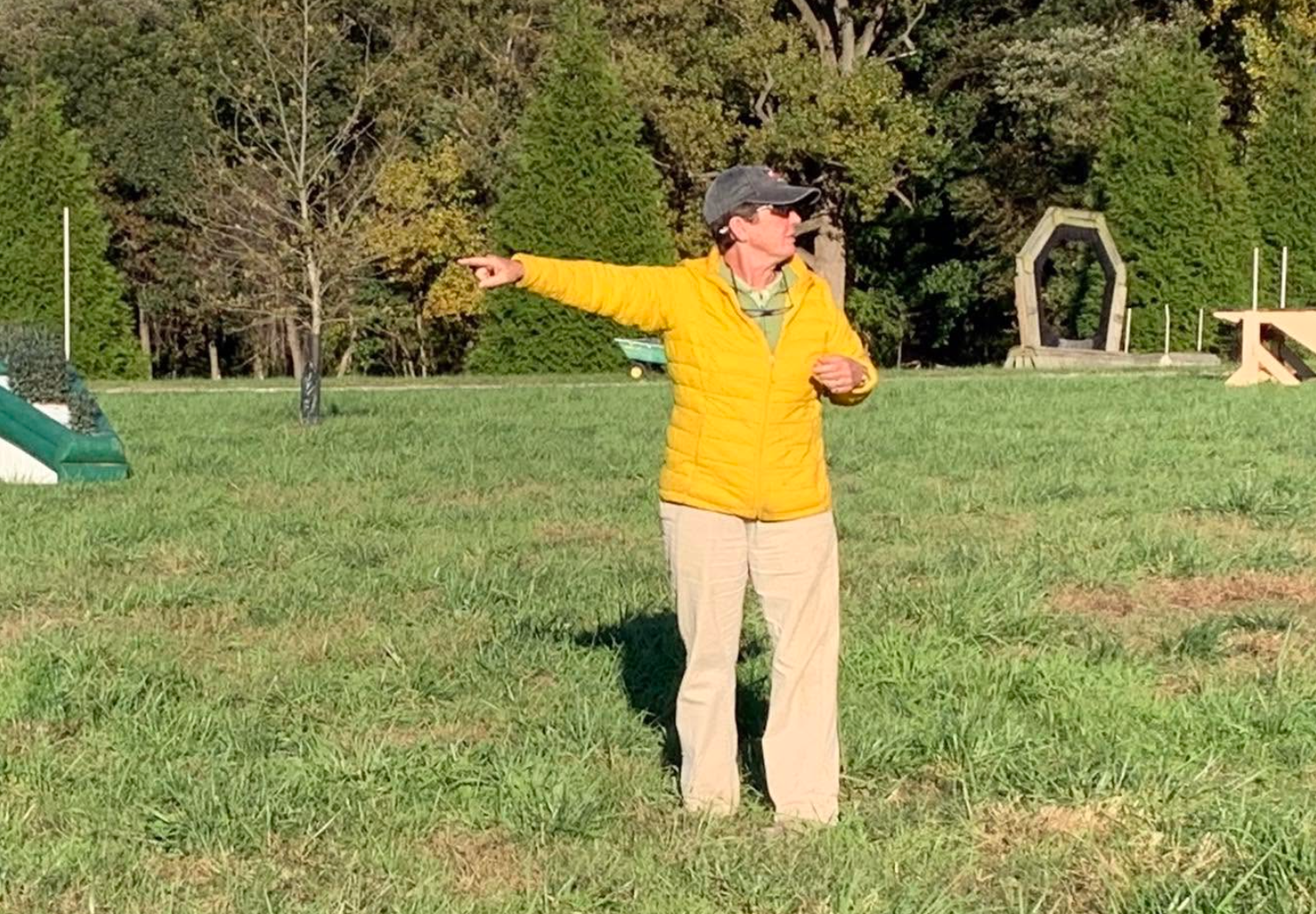 A man in a yellow jacket and beige pants standing in a grassy field, pointing to the left, with trees in the background.