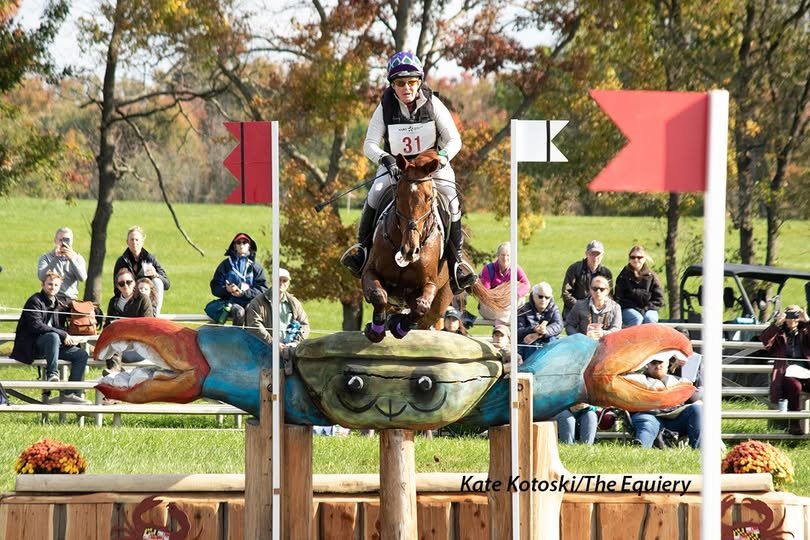 A female equestrian rider jumping over a crab-shaped obstacle during a horse jumping event, with spectators watching in the background.