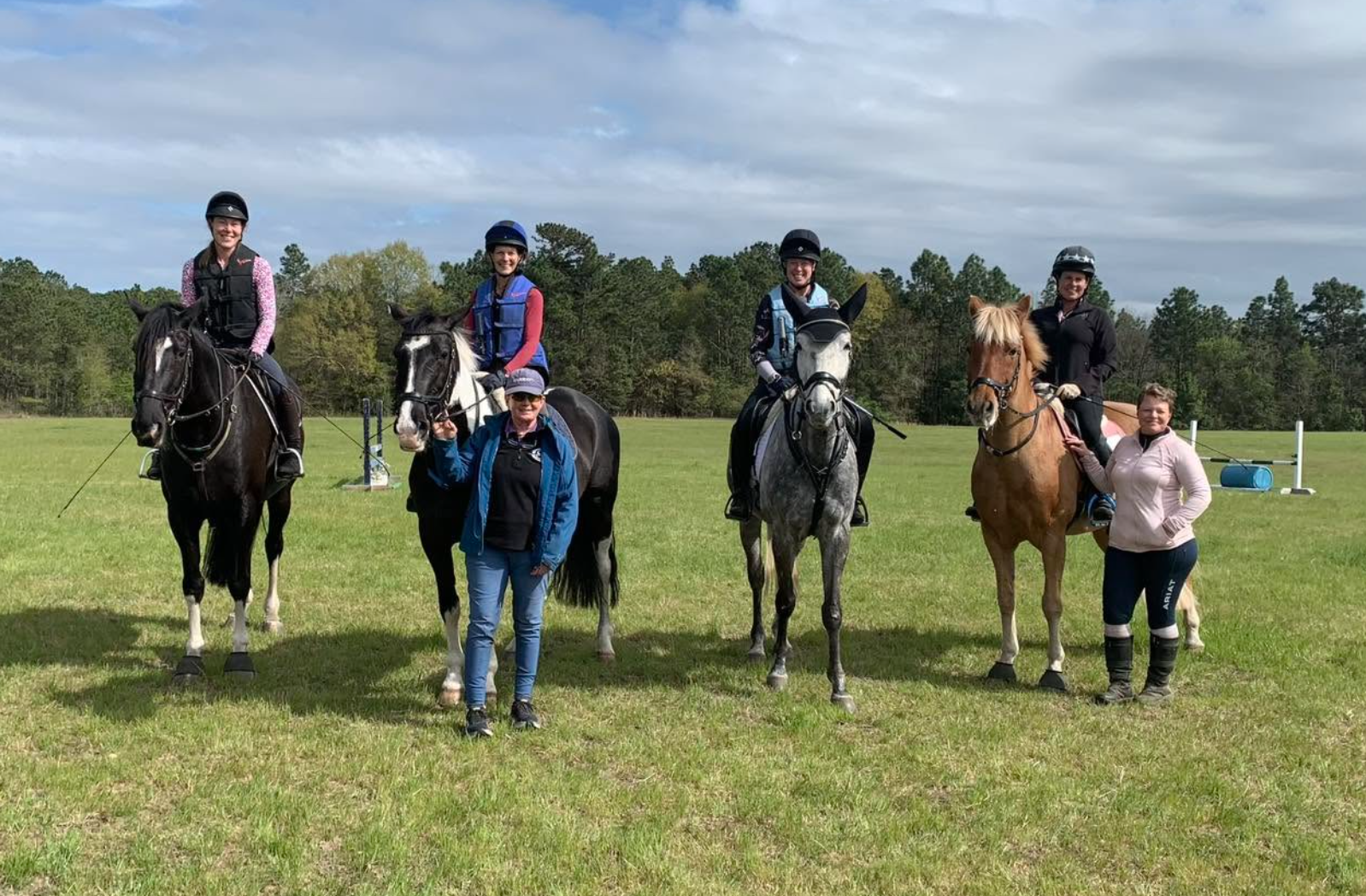 Group of five women and four horses in an open grassy field, with trees in the background. Three women are on horses, two women are standing beside horses, smiling at the camera. The women are dressed in riding gear, and the horses are equipped with riding saddles and bridles.