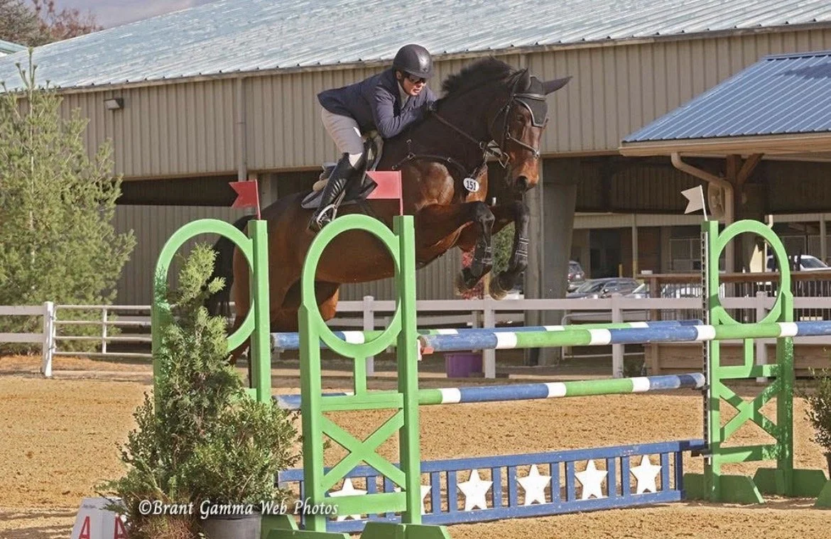 A person in riding gear and a helmet riding a brown horse over a colorful show jumping obstacle in an outdoor arena.