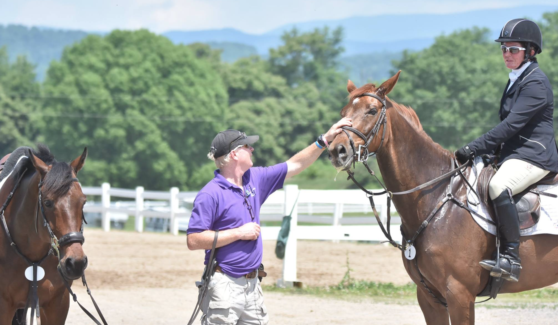A man in a purple shirt and black cap is petting a brown horse ridden by a person in riding attire, including a black helmet and sunglasses, on a sunny day at an outdoor equestrian facility with green trees and mountains in the background.