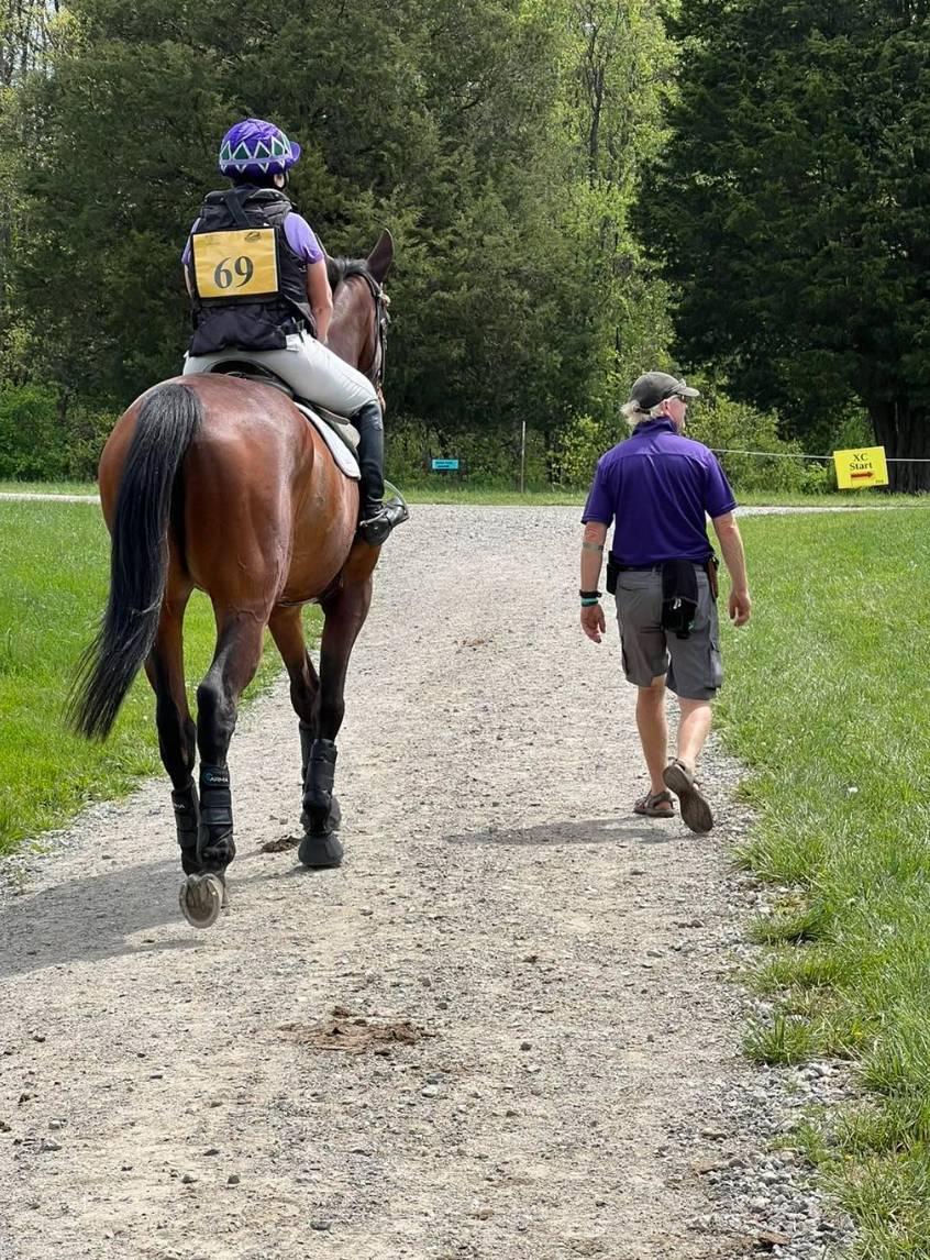 A person riding a horse with a number 69 riding bib, walking on a gravel path through a green, wooded area, with another person walking beside the horse, wearing a purple shirt and gray shorts.