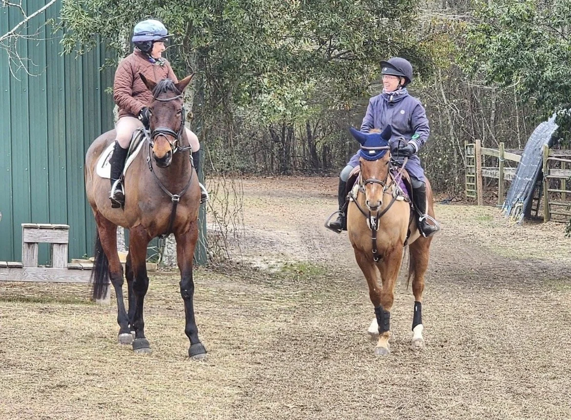 Two people riding horses on a dirt path, surrounded by trees and bushes, with a green building on the left and some equipment on the right.