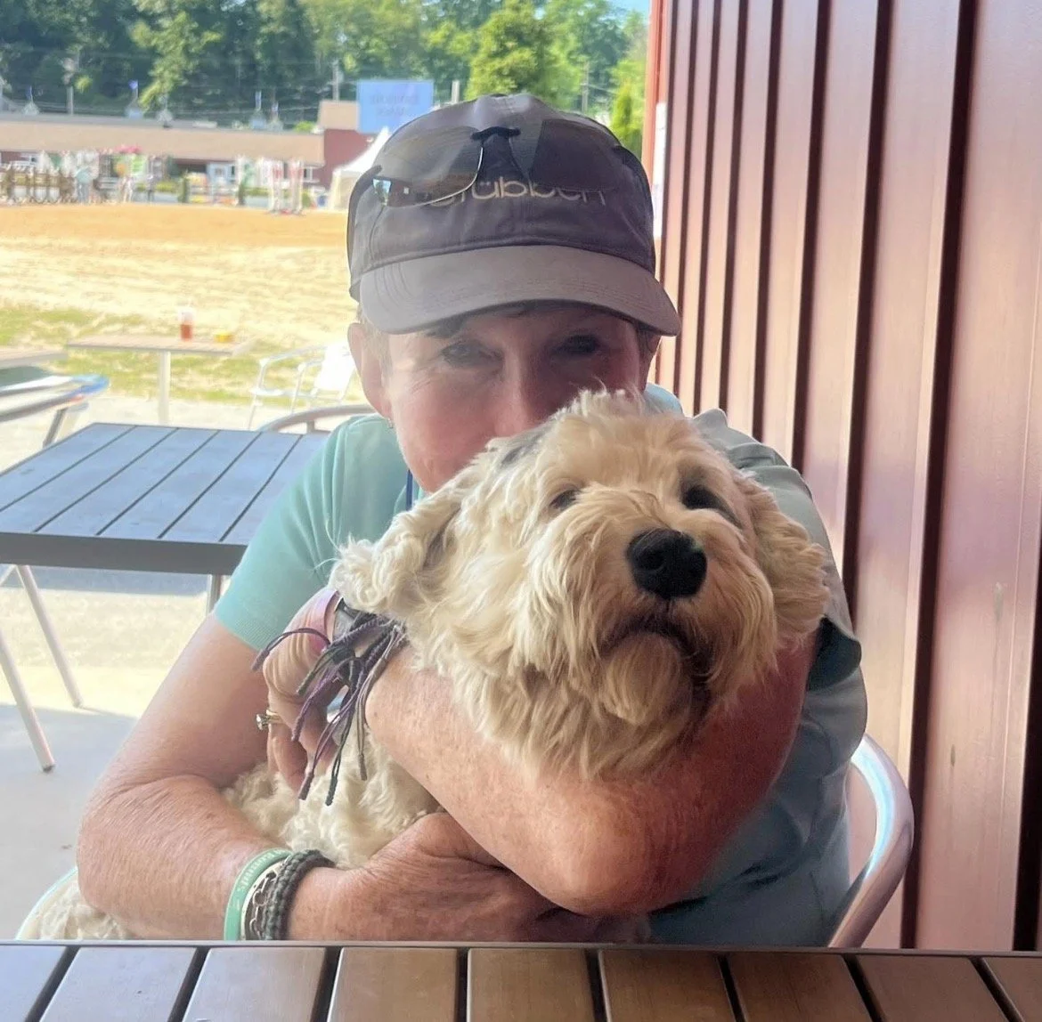 A woman holding a golden doodle dog inside a restaurant or cafe, with outdoor seating and a field visible through the window in the background.