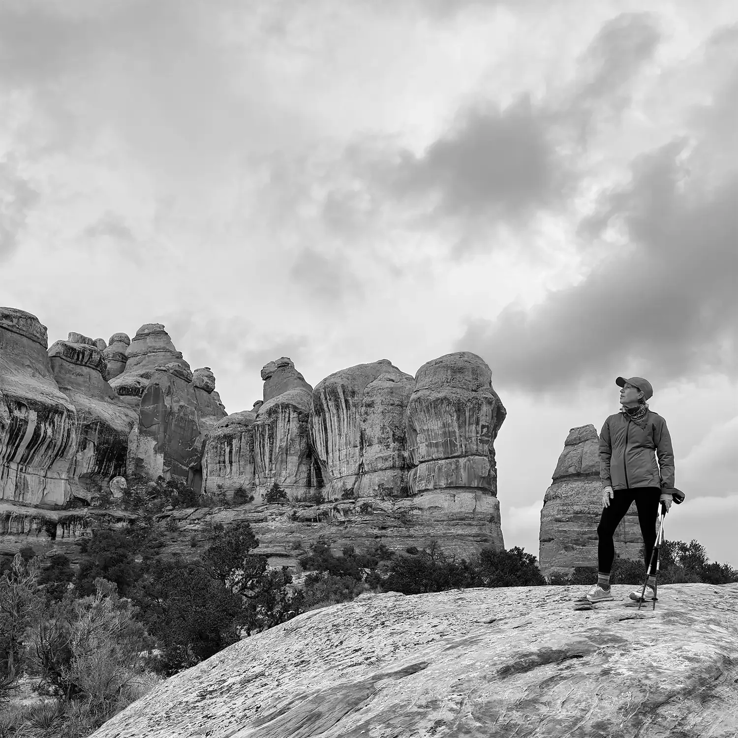 Dagny Scott -  a woman with a walking stick standing on a rocky terrain with large rock formations and a cloudy sky in the background.