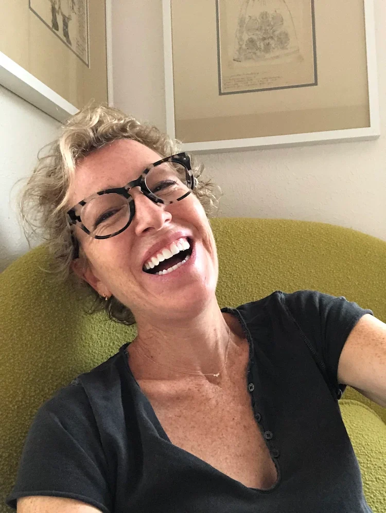 Dagny Scott - a woman with curly blonde hair, wearing glasses and a black shirt, smiling and sitting on a yellow chair, with framed artwork on the wall behind her.