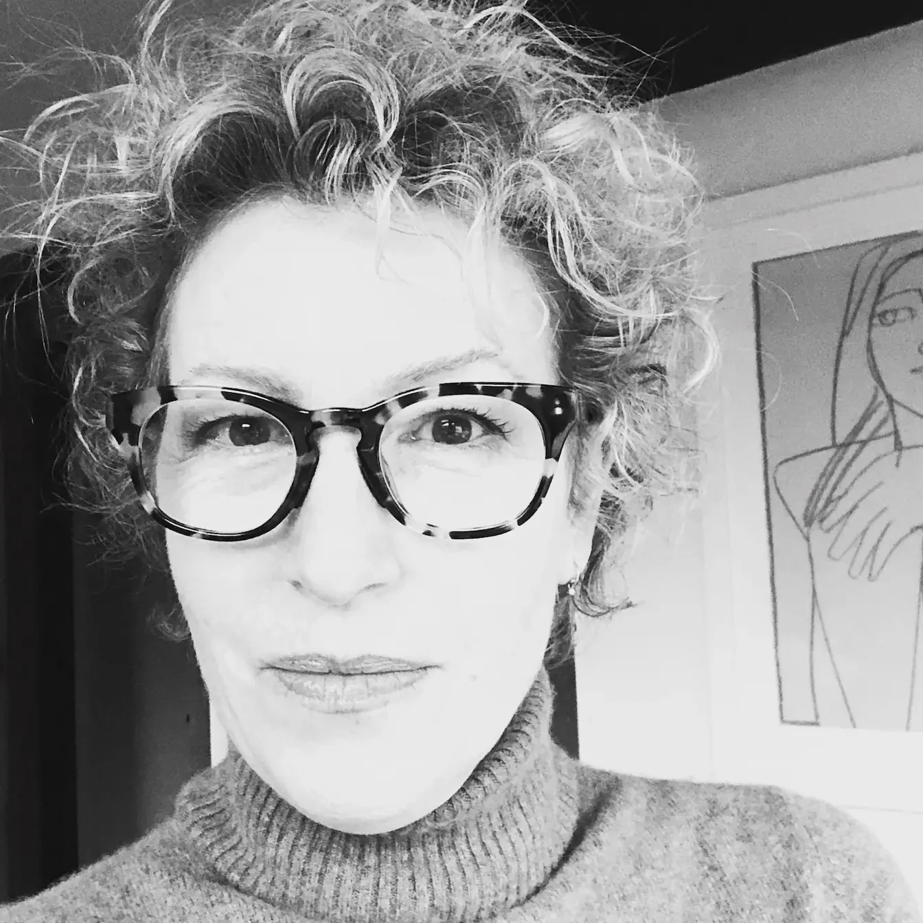 Dagny Scott - a woman with curly hair wearing glasses and a turtleneck sweater, looking slightly upward in a black and white photo, with a framed line drawing of a person in the background.
