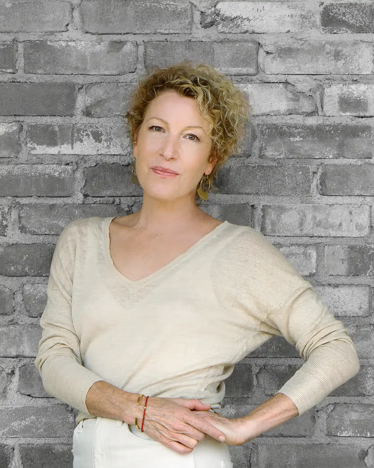 Dagny Scott - a woman with curly blonde hair, wearing a beige V-neck sweater and gold earrings, standing against a gray brick wall, looking at the camera with a confident expression.