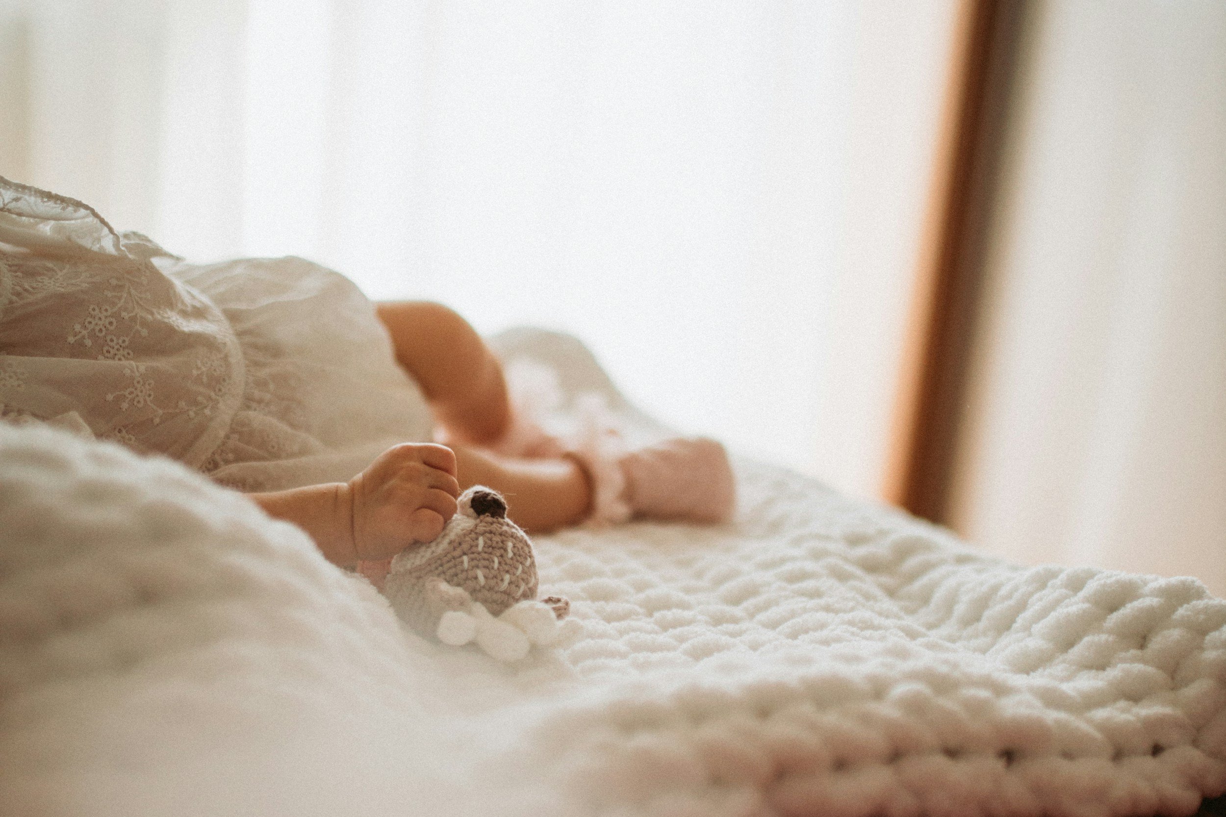 A baby lying on a bed covered with a knit blanket, wearing pink clothing and holding a small crochet toy.
