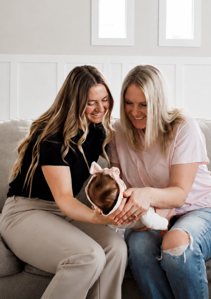 Two women hold a baby girl in a pink headband with a bow, sitting on a gray couch in a bright room with white walls and picture frames. The women are smiling and looking at the baby.