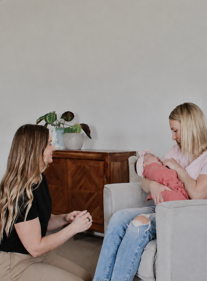 A woman holding a baby and smiling at her, while another woman kneels in front of them, smiling and holding her hands together.