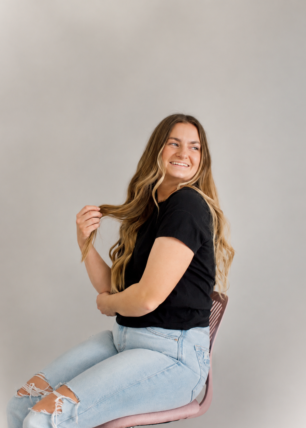 Young woman with long wavy hair sitting on a pink chair, smiling and looking to the side against a plain gray background.