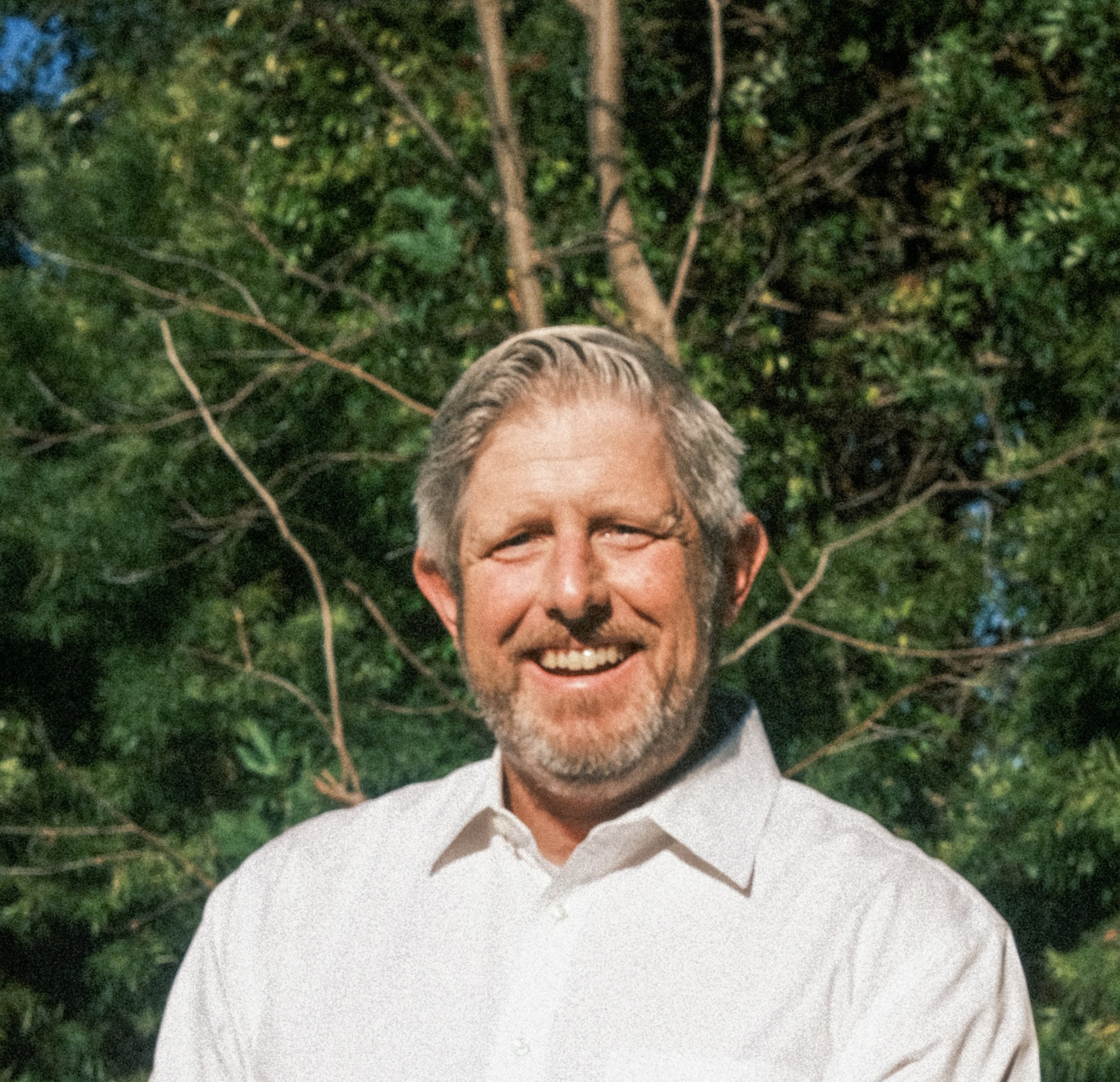A man with gray hair and a beard smiling, outdoors with green trees in the background.