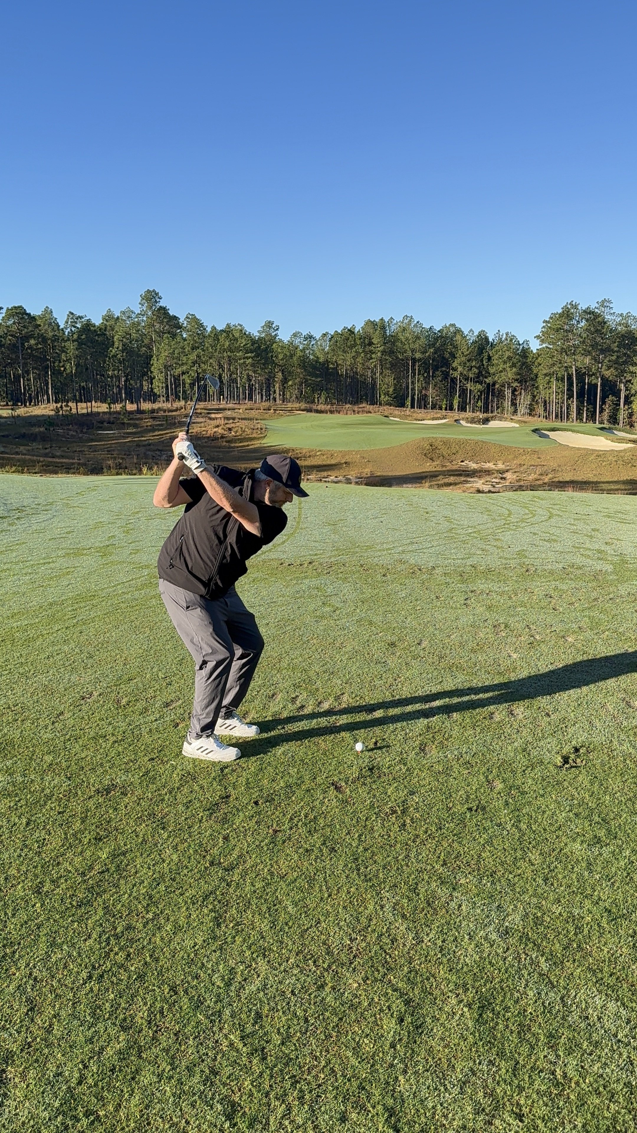 A man in a black jacket and gray pants playing golf on a green golf course under a clear blue sky. He is in the process of swinging a golf club, preparing to hit the golf ball.