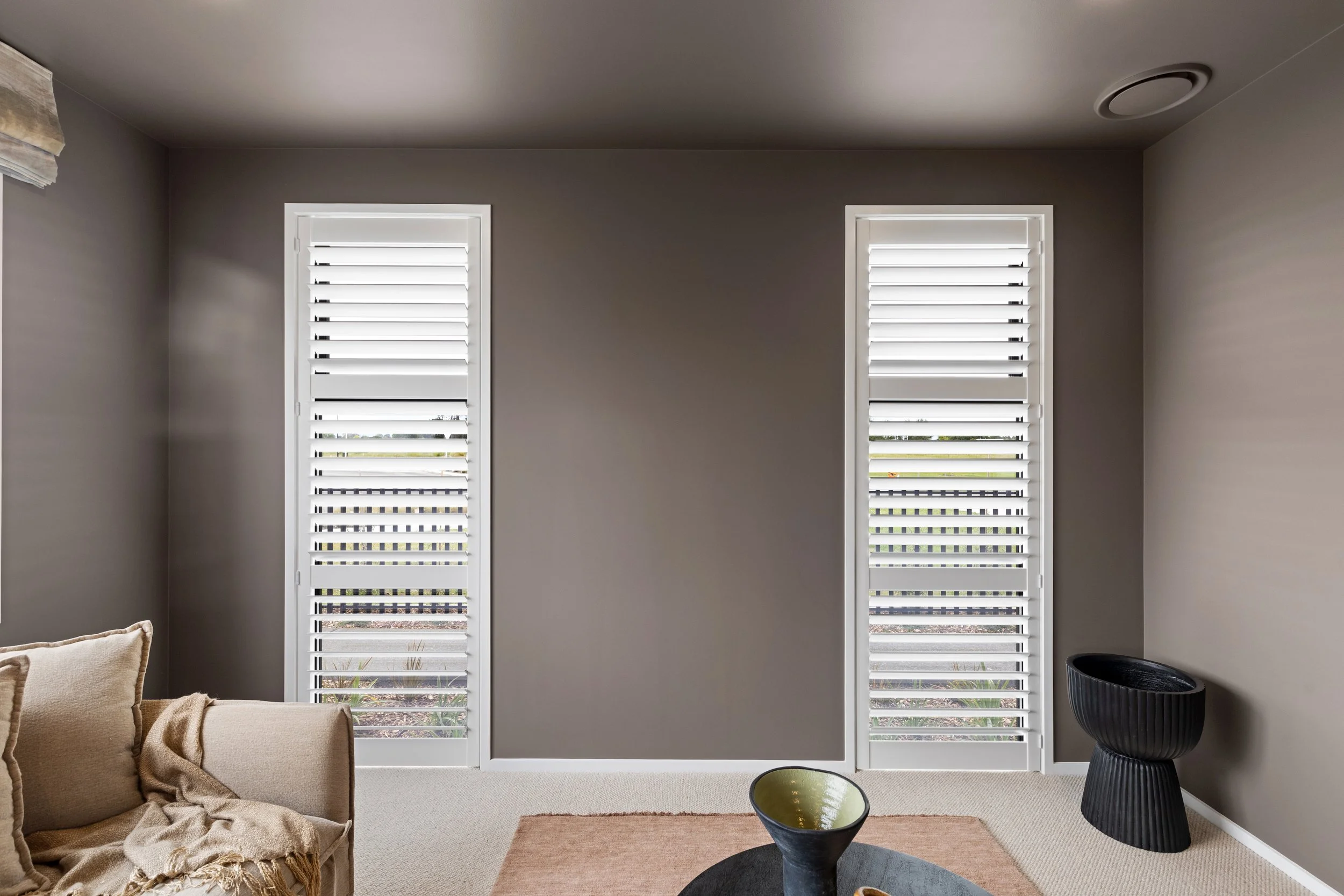 Living room with beige sofa, beige throw blanket, and pillows, beige carpet, brown and black coffee tables, two tall white plantation shutters on windows, gray walls, and decorative black side table.