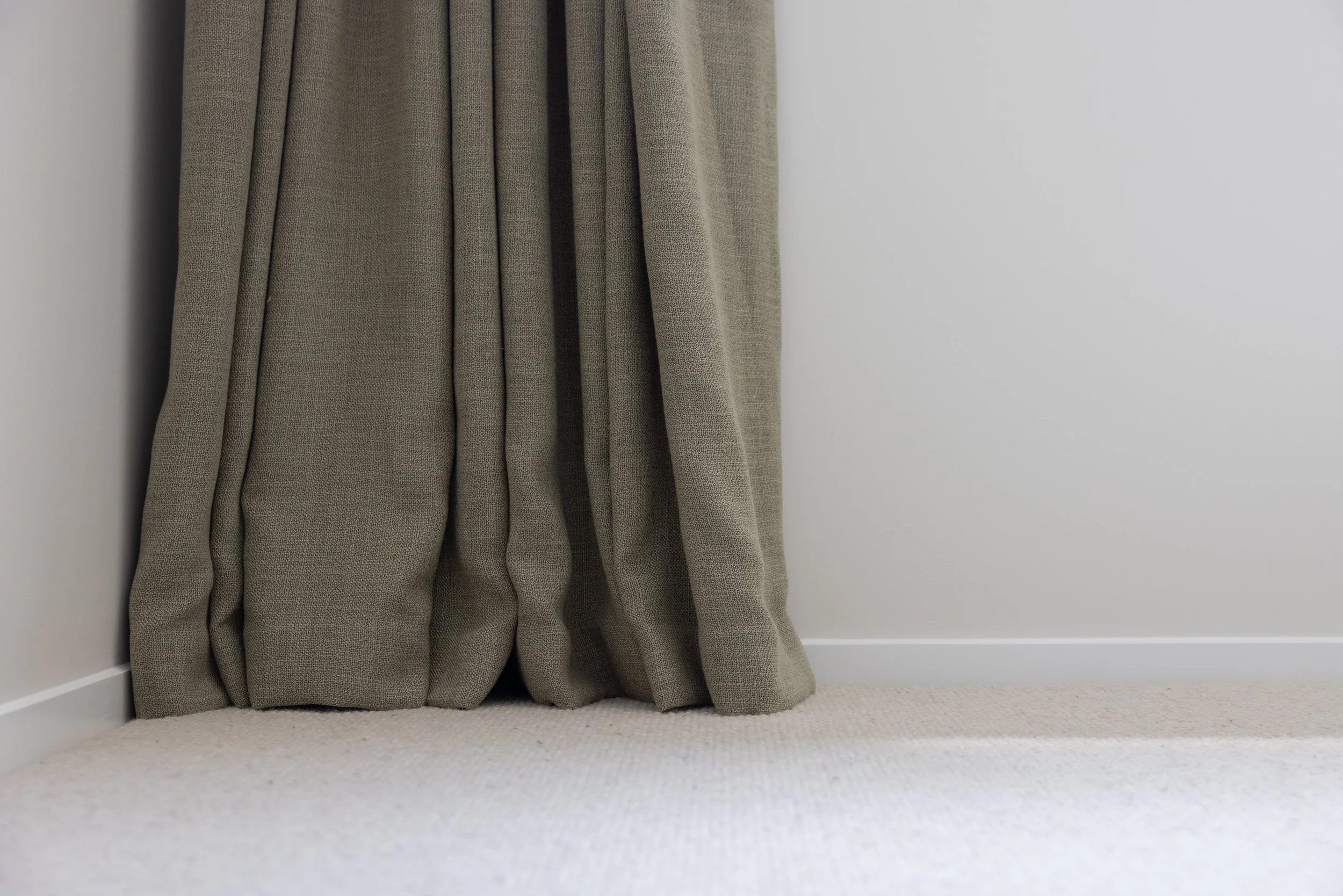 Close-up image of beige curtains hanging near a white wall and light-colored carpeted floor.