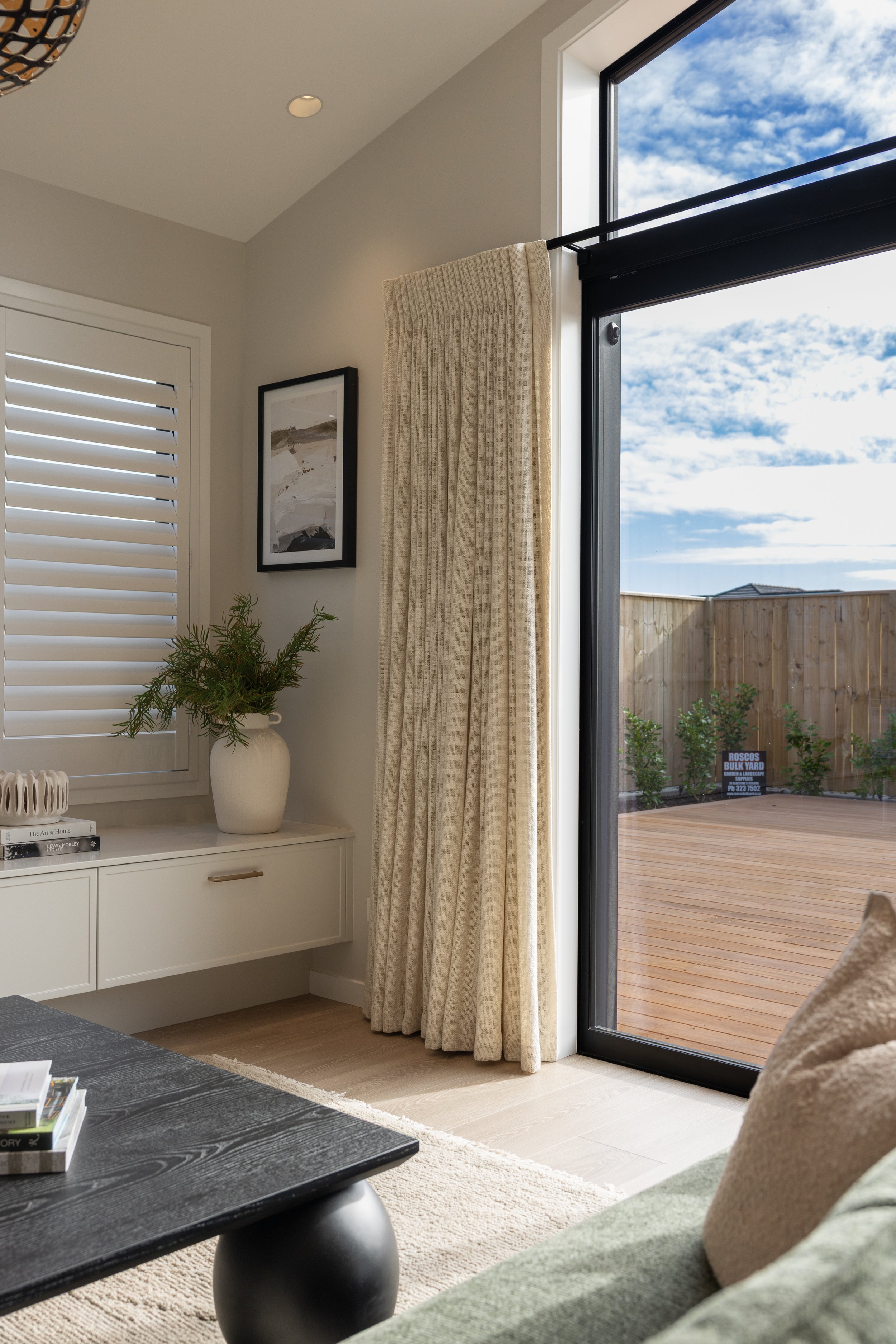 Interior view of a living room with a white cabinet and potted plant next to a window with cream curtains. Outside, a wooden deck and a cloudy sky are visible.