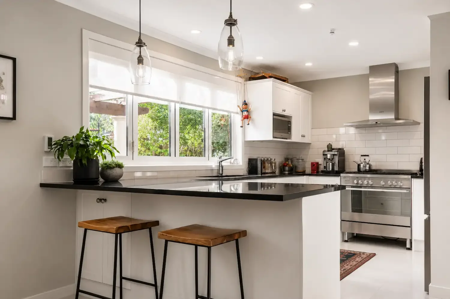 Modern kitchen with white cabinets, black countertop, window with greenery outside, plants on the counter, and two wooden stools at the breakfast bar.