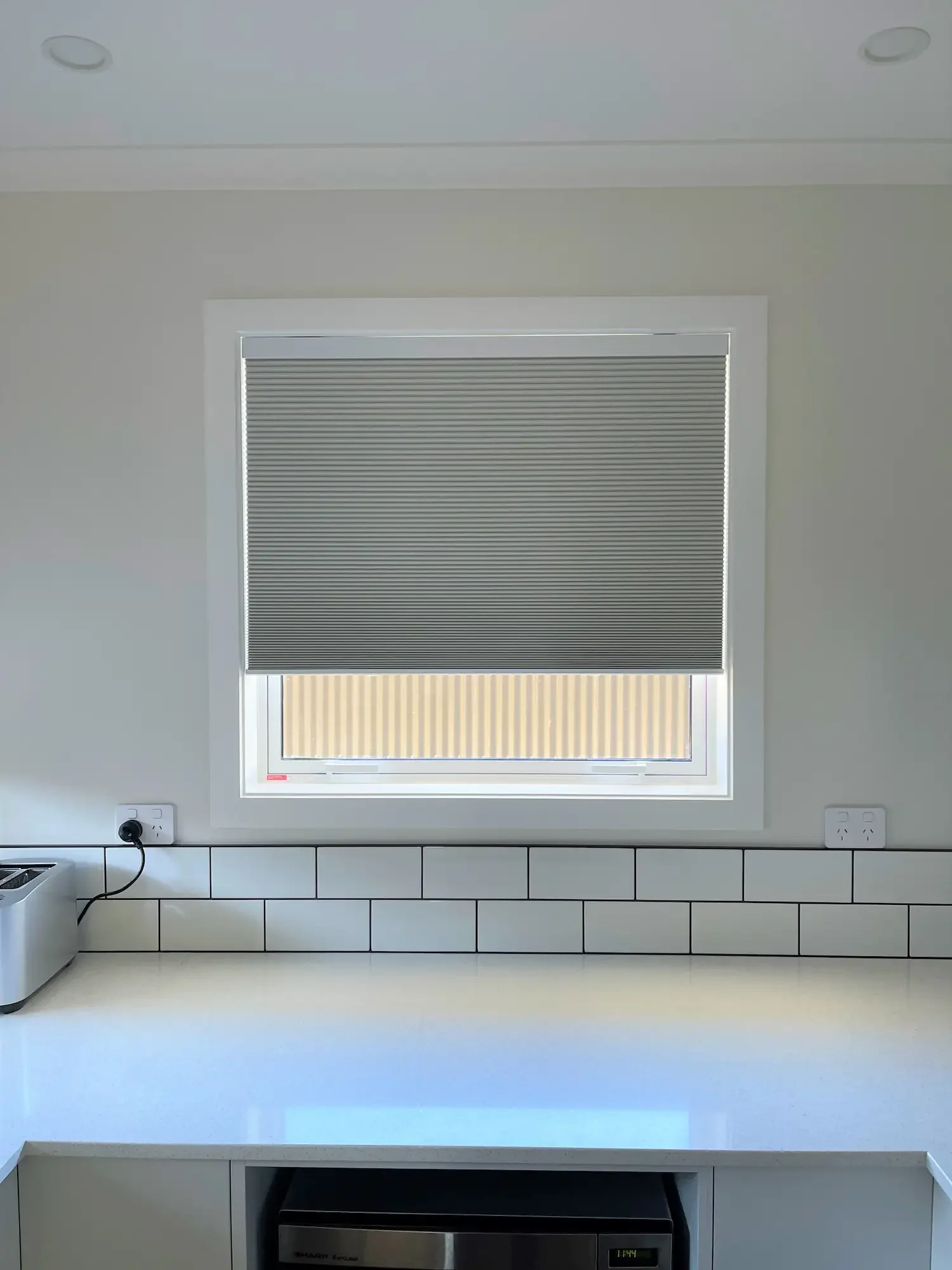 A window with a closed grey pleated blind, framed in white, on a white wall in a modern kitchen with white tiled backsplash and electrical outlets.