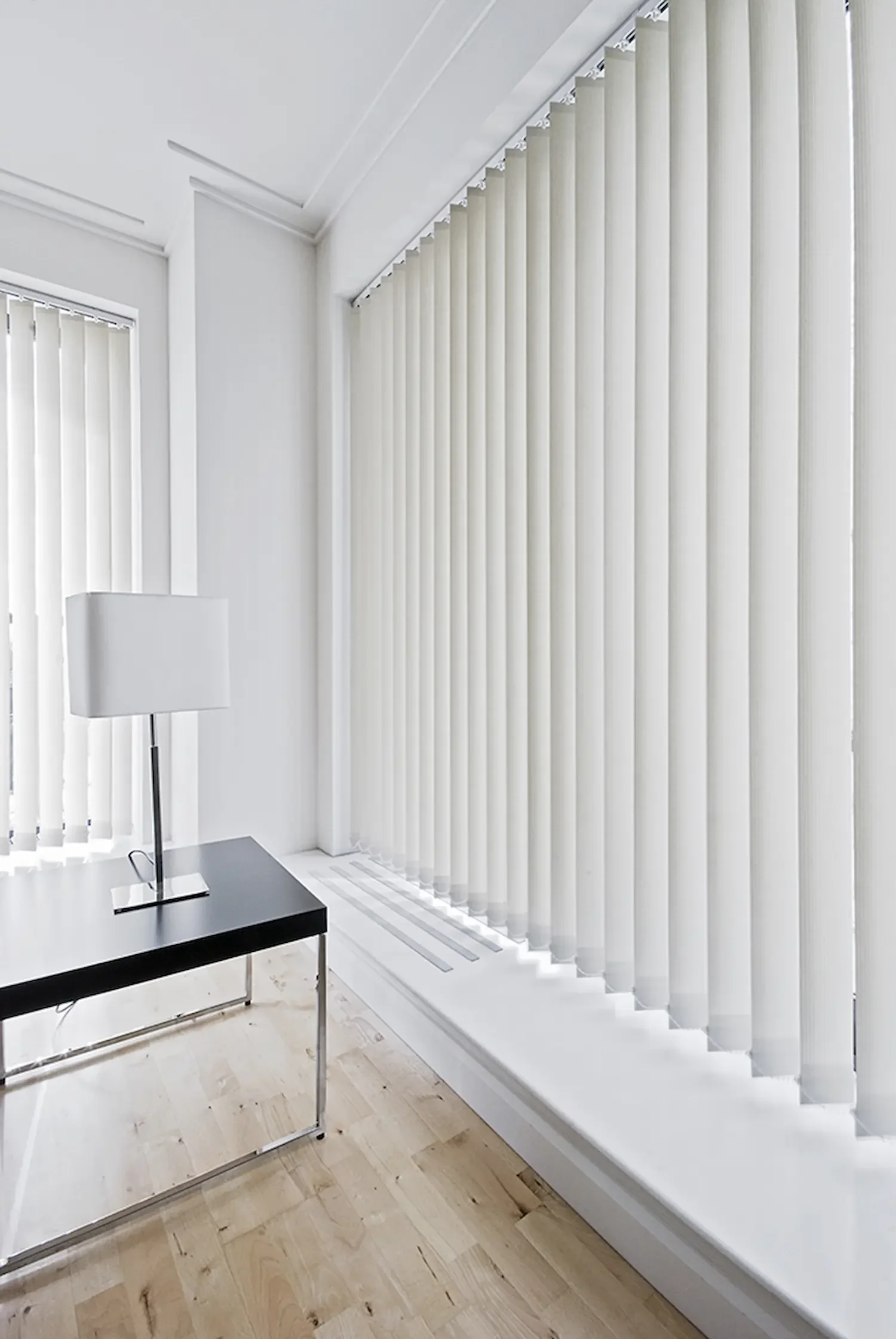A corner of a room with white walls and vertical blinds covering large windows; a black coffee table with a silver and white table lamp on it is in the foreground on a light wood floor.