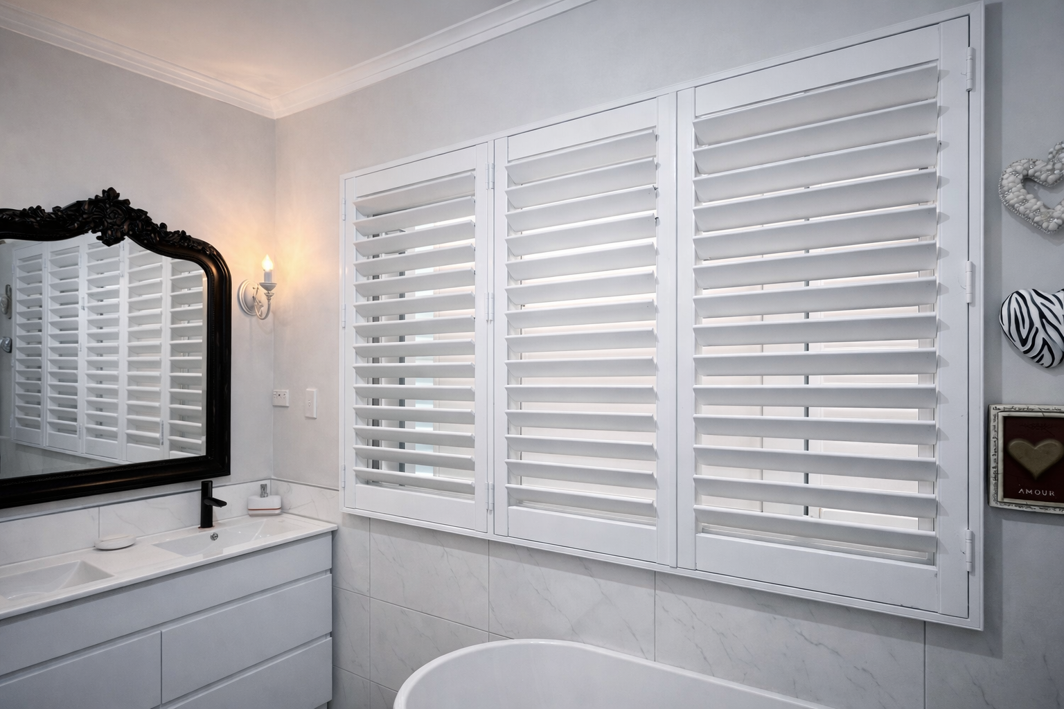 Bathroom with white shutters on window, black-framed mirror, white sink with black faucet, and wall decorations including a zebra-patterned hat and a heart-shaped wall art with the word 'amour'.
