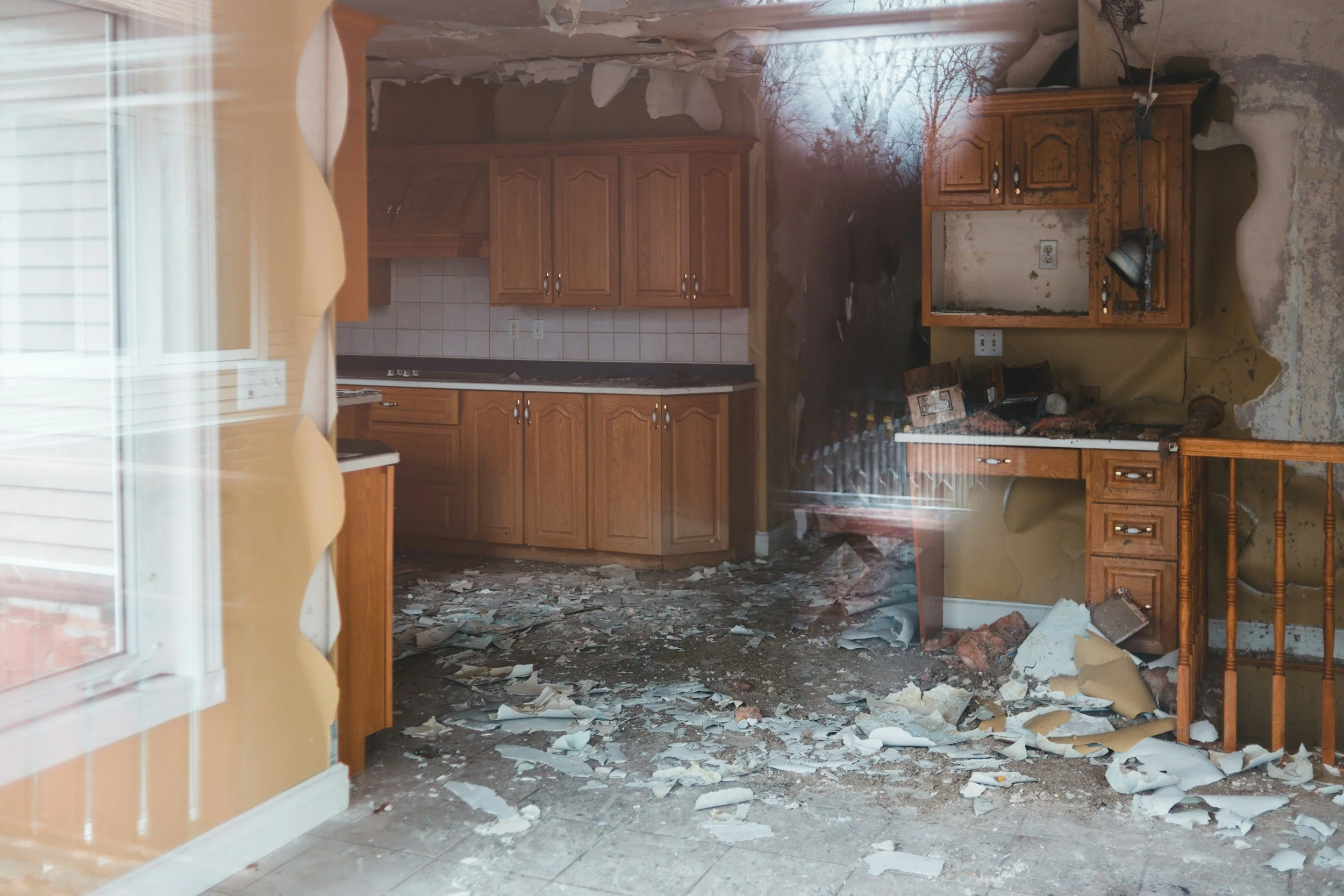 A kitchen viewed through a window with yellow curtains. The kitchen is damaged with debris, broken ceiling, and wall damage. Wooden cabinets and a tiled backsplash are visible, alongside scattered debris and damaged surfaces.