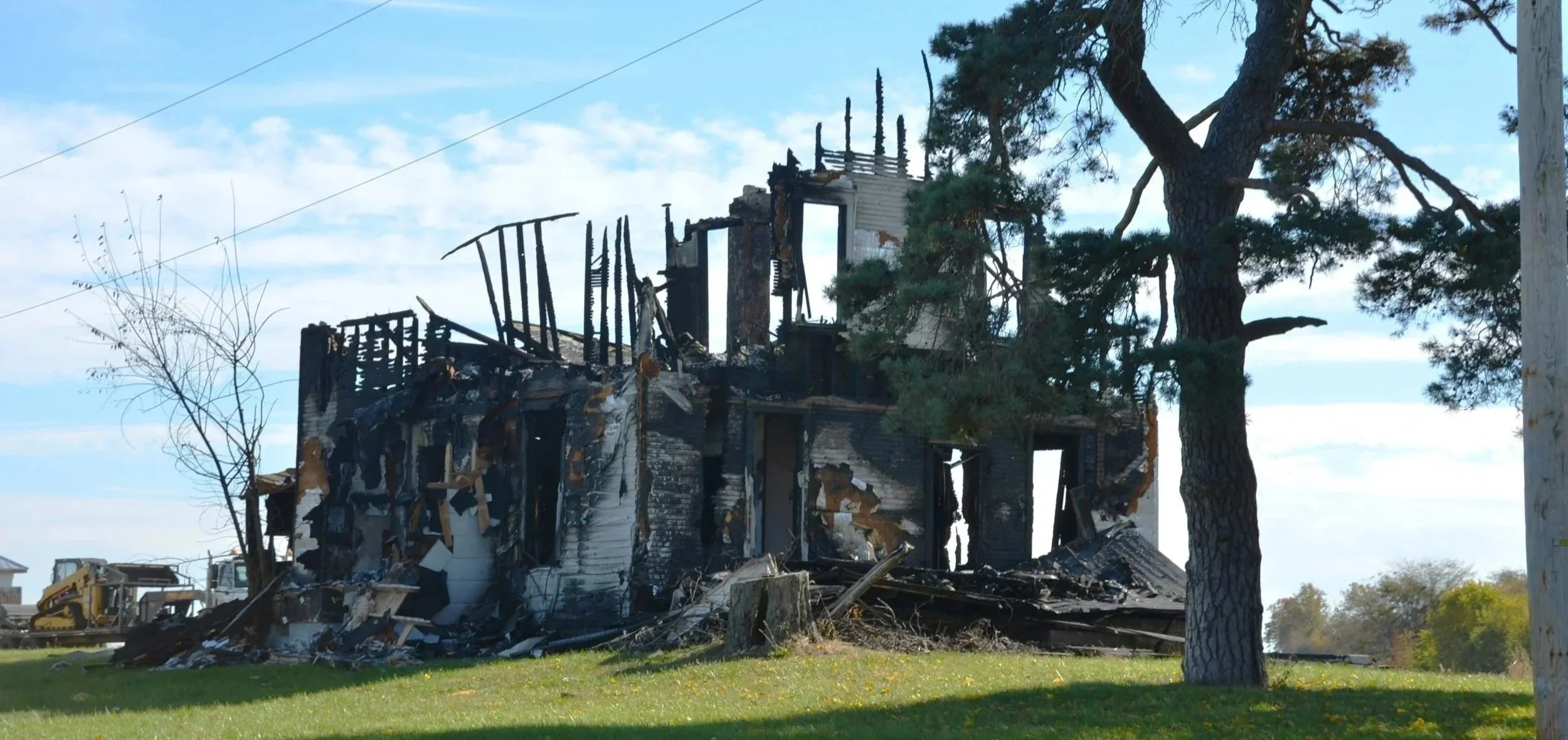 A house heavily damaged by fire, with charred walls and a partially collapsed roof, surrounded by green grass and some trees.