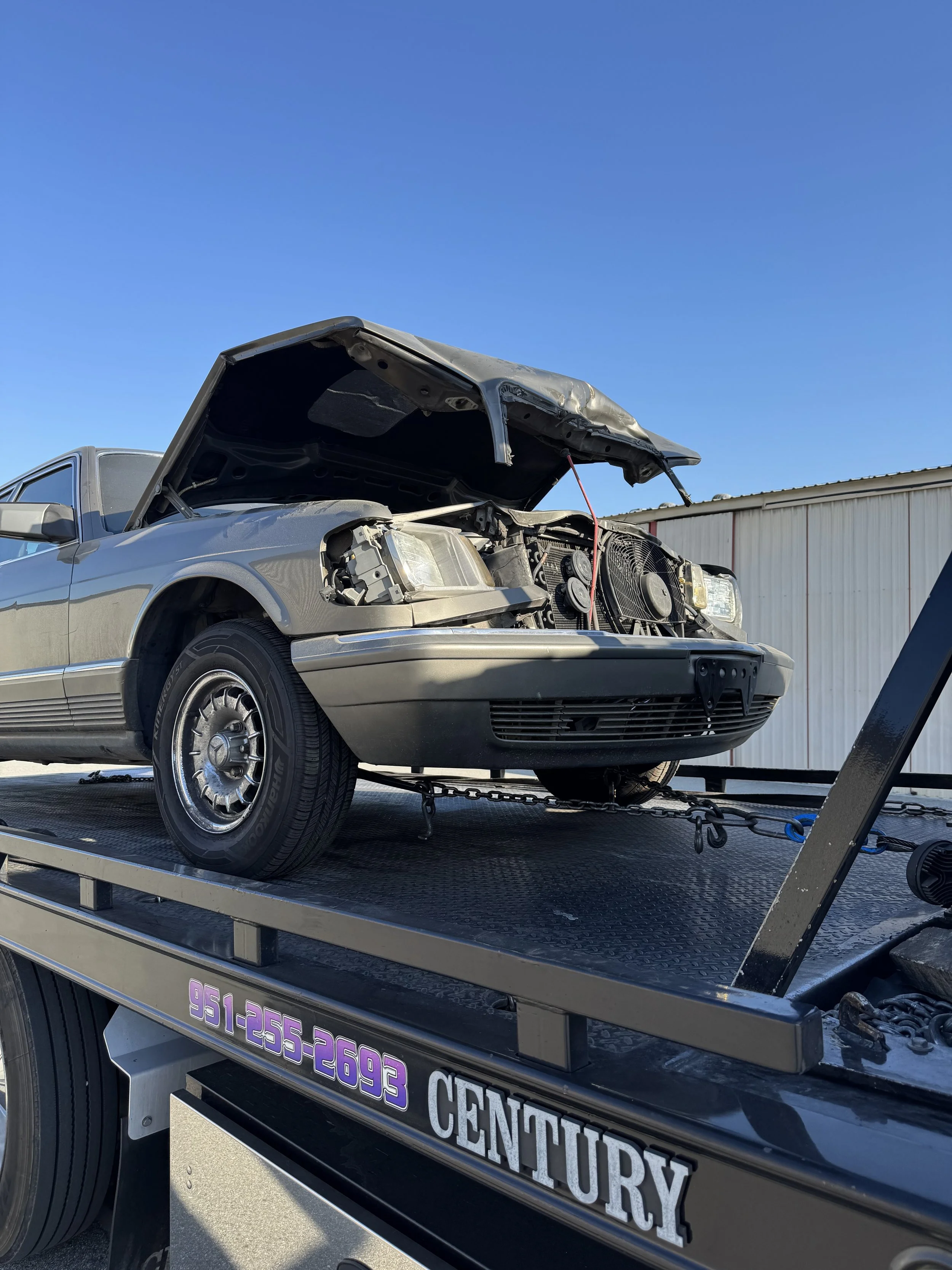 Silver car with hood open on tow truck, at a repair shop on a sunny day.
