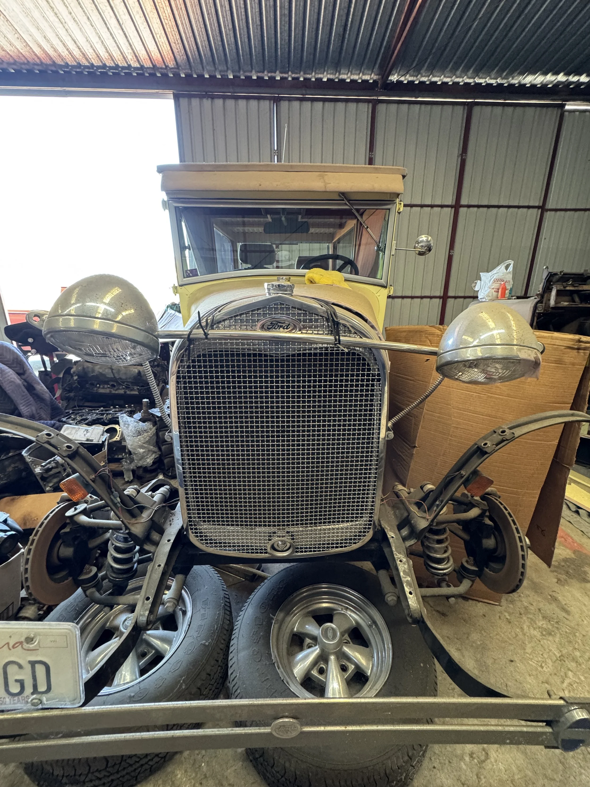 An old yellow vintage Ford car with round headlights underneath a metal roof, surrounded by tires and equipment in a storage or workshop setting.