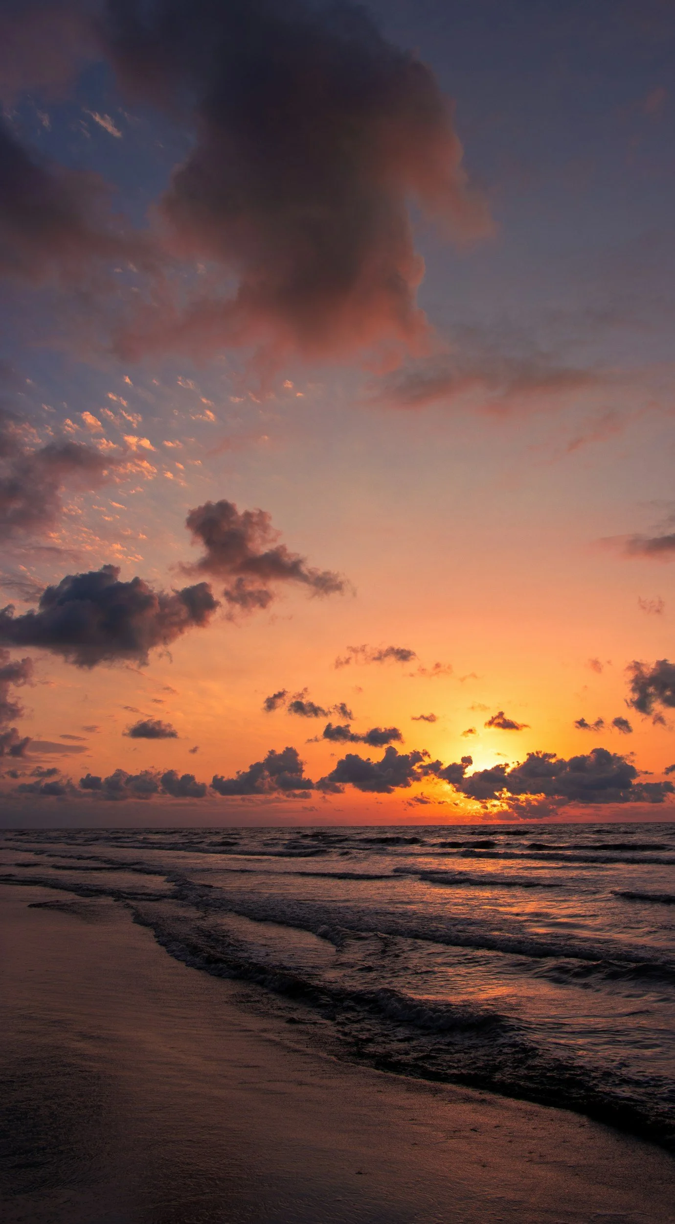 Sunset over the ocean with colorful clouds in the sky and waves crashing onto the sandy shore.